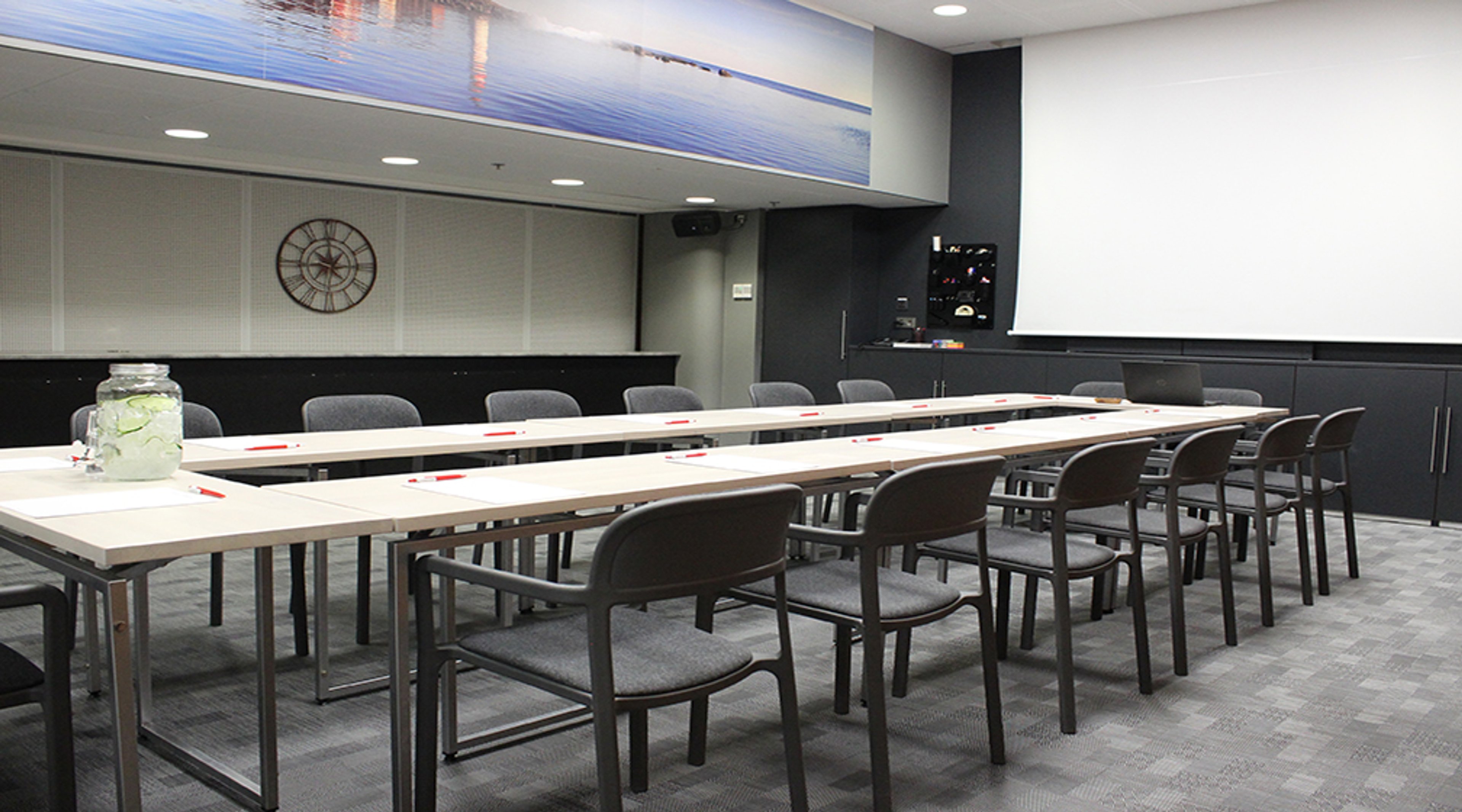 Moderni kokoushuone, jossa pitkät pöydät ja harmaat tuolit ympärillä. Seinällä on kello ja seinävalokuva veden äärellä.