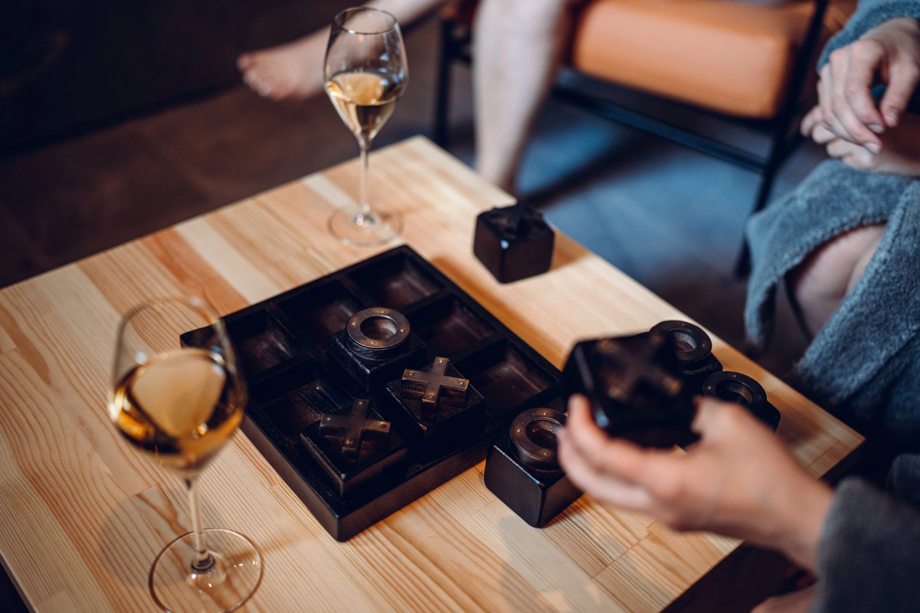 Puinen pöytä, jolla on ristinolla-peli mustilla paloilla ja kaksi lasia valkoviiniä. Henkilö pitelee pelipalaa.