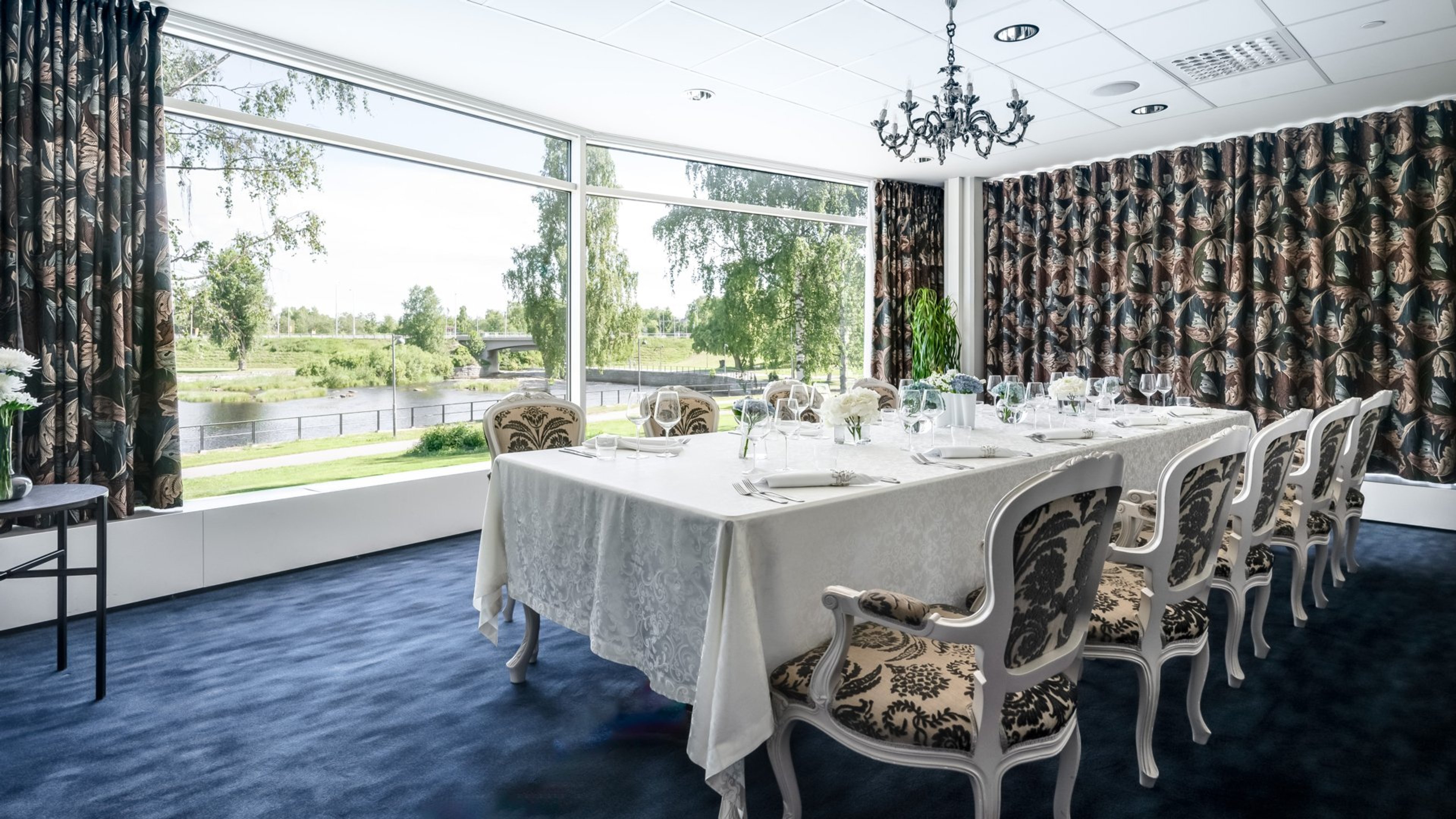 Juhlahuone, jossa pitkä pöytä valkoisen pöytäliinan ja kattauksen kera. Koristeelliset tuolit, isot ikkunat joista näkyy joki ja puistomaisema.