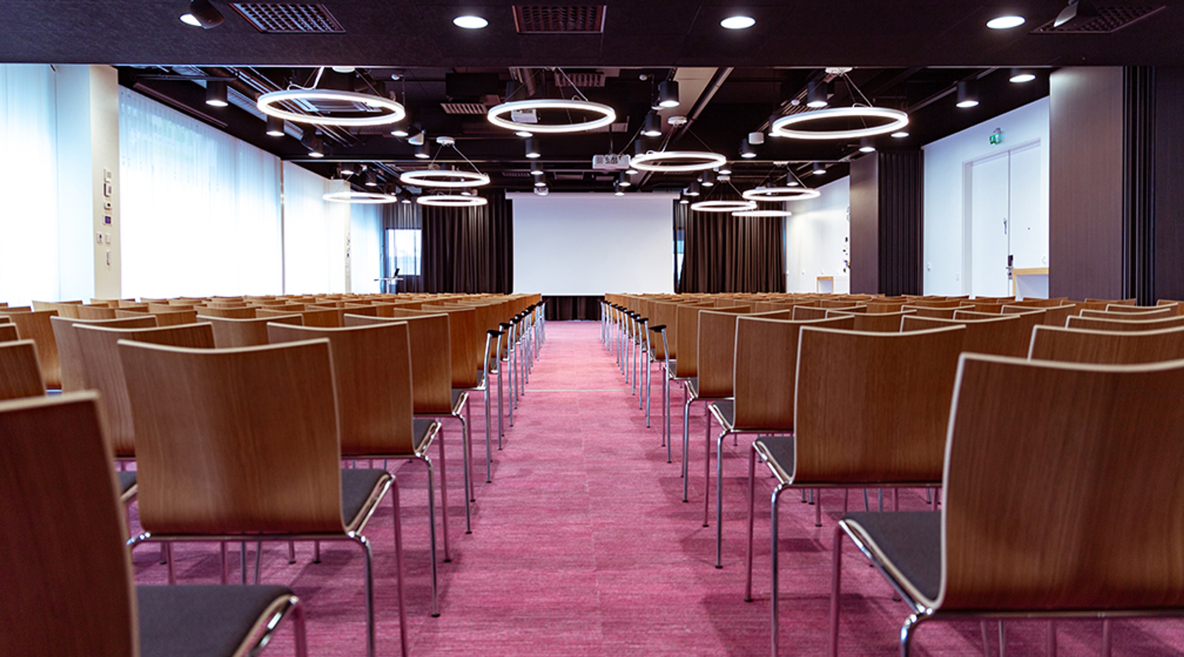 Modern auditorium with wooden chairs and red carpet. Ceiling features circular lights and a white projection screen at the front.