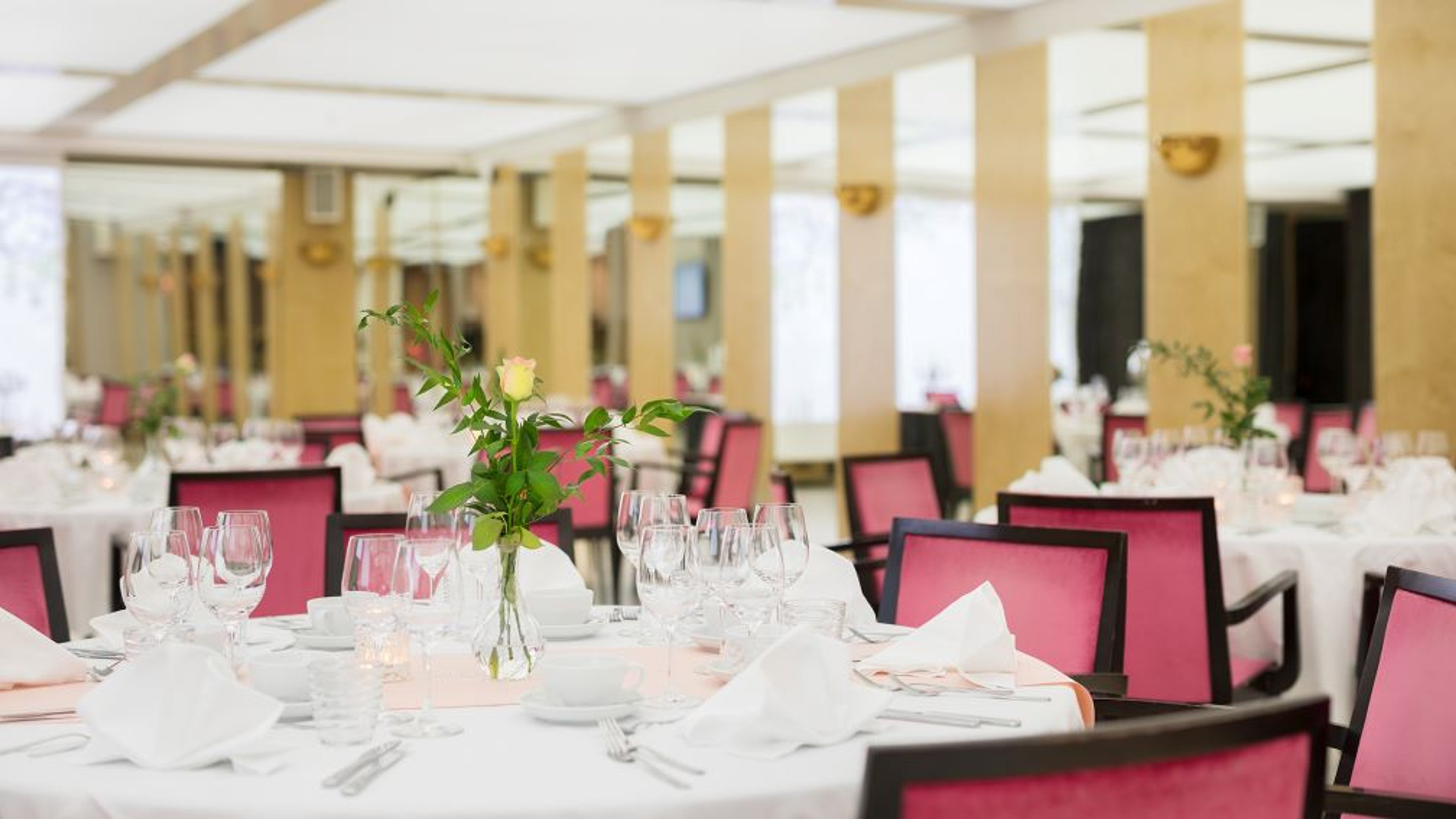 Elegantti ruokasali, jossa on pyöreitä pöytiä valkoisilla liinoilla, pinkillä verhoiltuja tuoleja ja pienet kukka-asetelmat pöydillä.