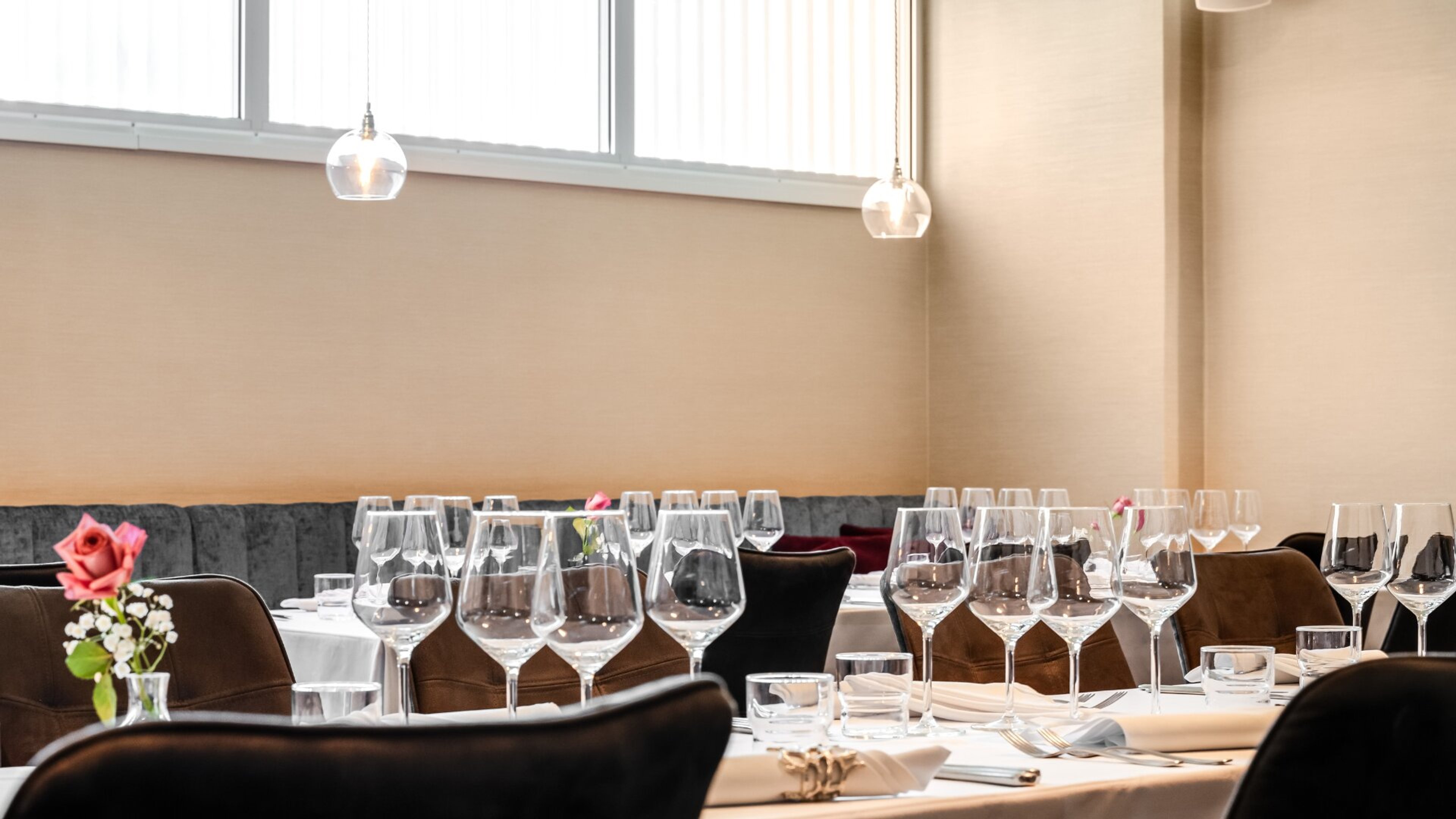 Tyylikkäästi katetut kabinetin pöydät viinilaseineen, harmaa sohvaistuin seinän vierellä ja pienet kukka-asetelmat pöydillä.