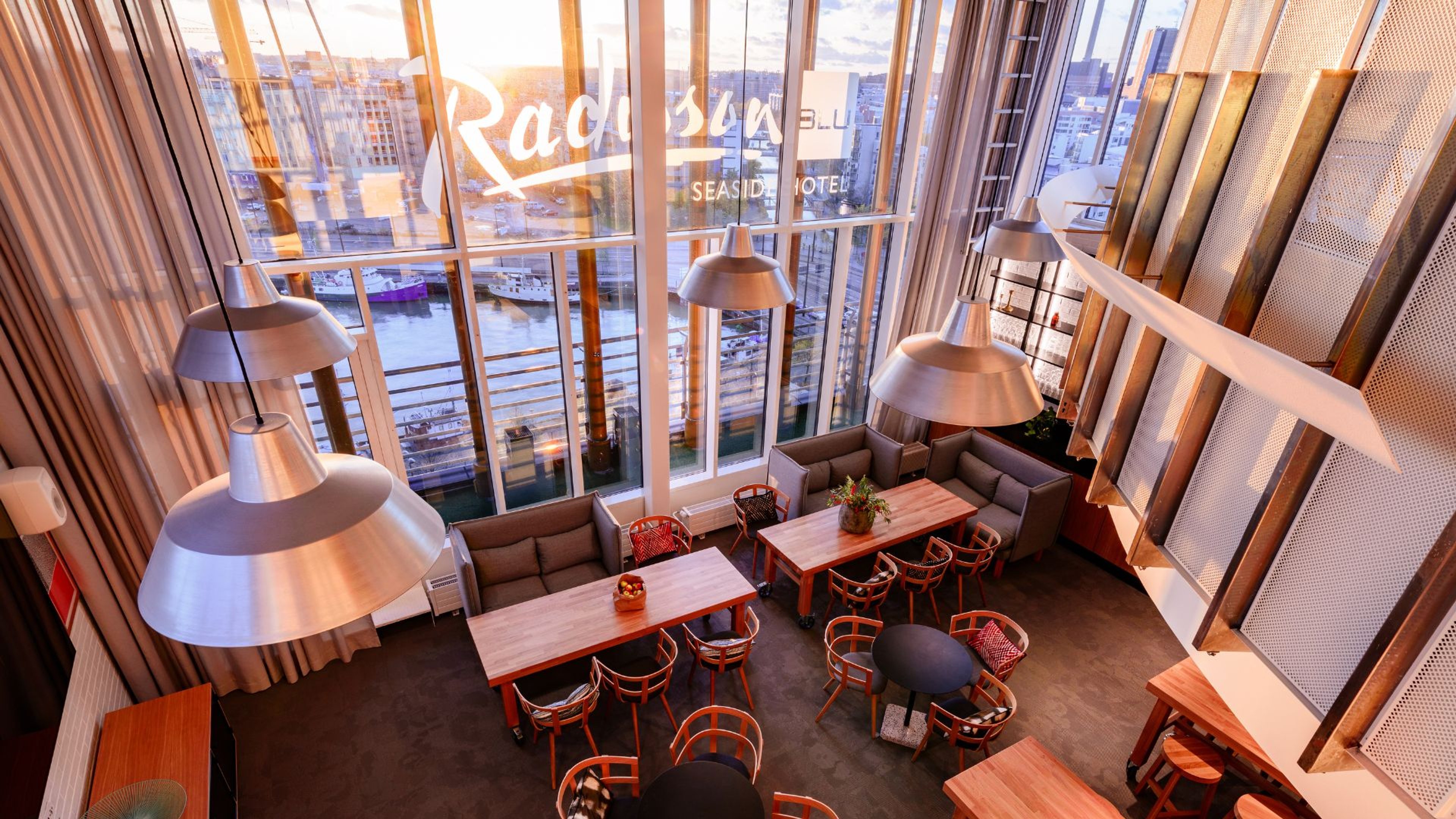 Hotellin lounge-alue isoilla ikkunoilla ja näkymällä merelle, puiset pöydät, tuolit ja sohvat, tasainen betonilattia.