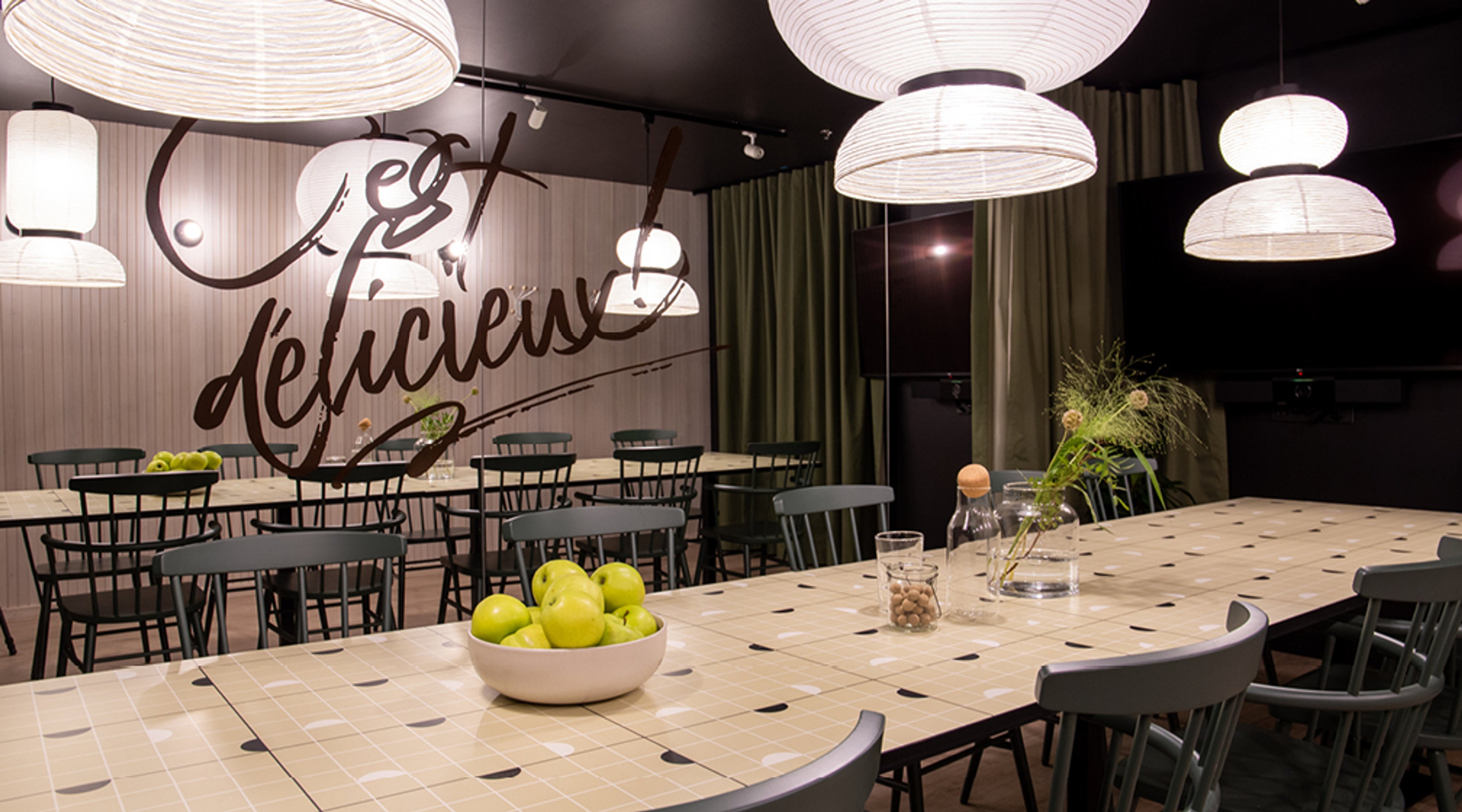 Moderni ruokailutila, jossa vaaleat riippuvalaisimet, kaksi ruokapöytää, vihreitä tuoleja ja kulho vihreitä omenia pöydällä.