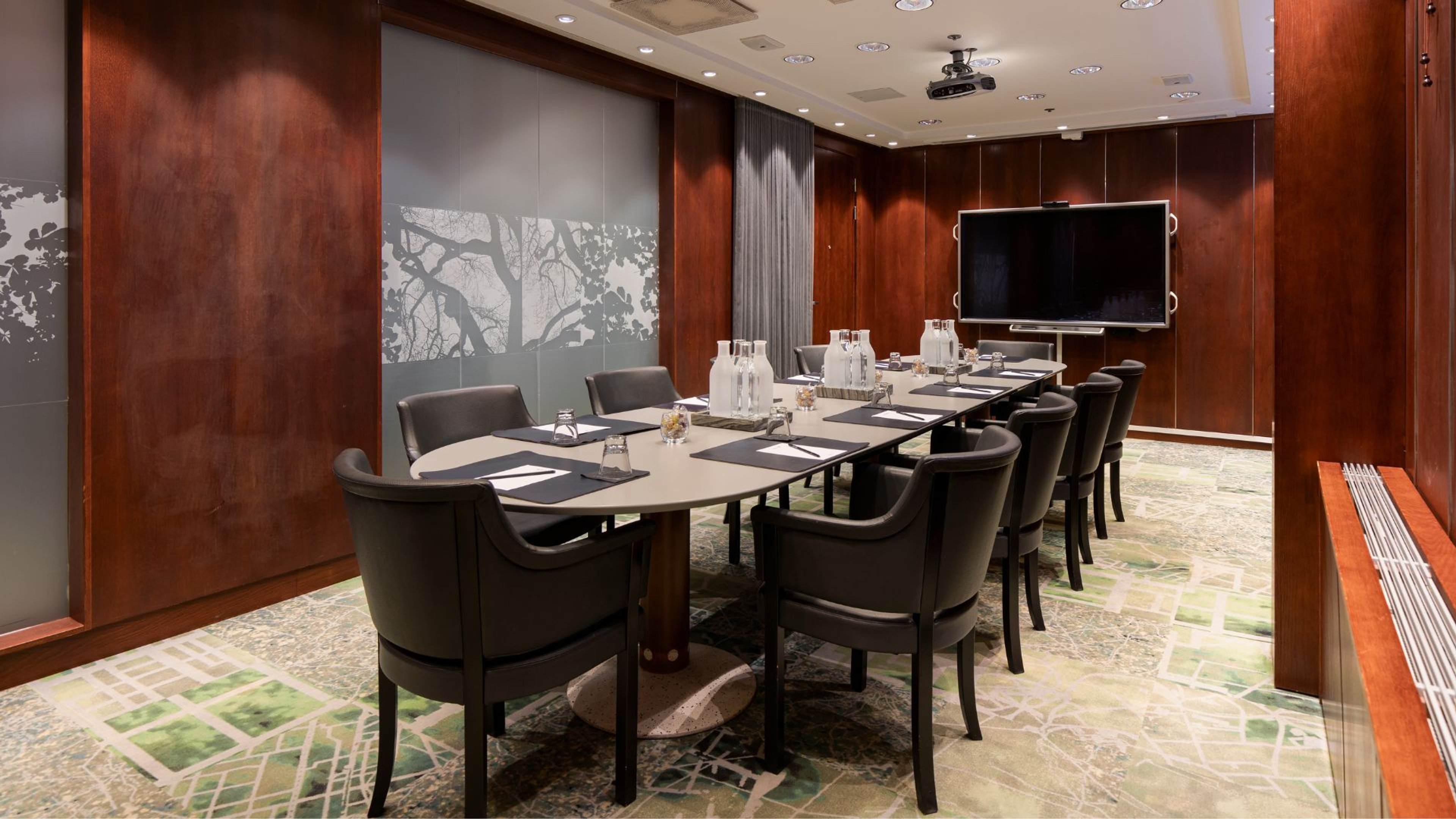 Modernissa kokoushuoneessa on ovaalinmuotoinen pöytä, tummat tuolit, puupaneelit ja TV. Huoneessa on hyvä valaistus ja tasainen lattiapinta.