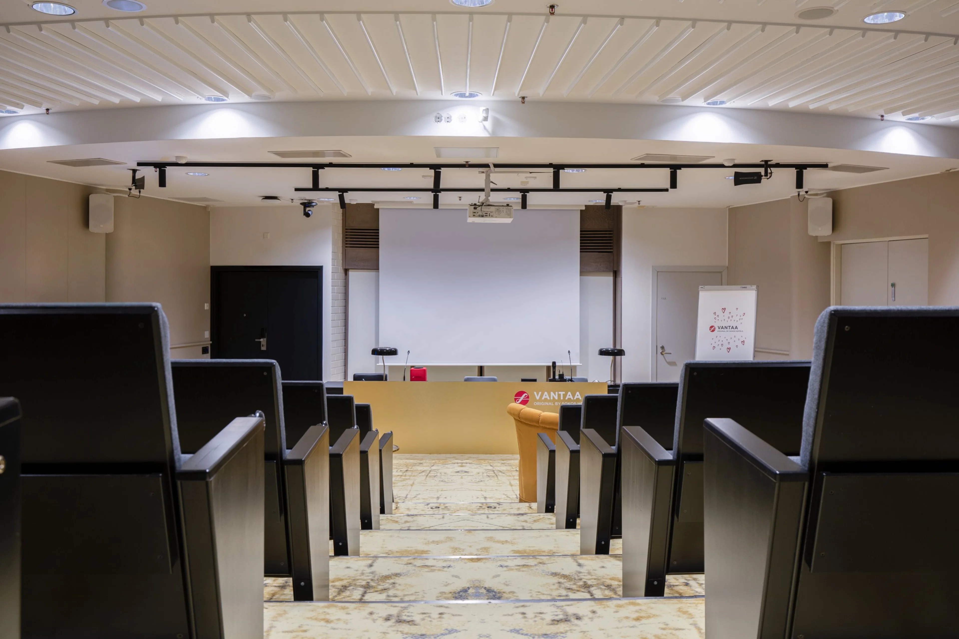 Small auditorium with tiered seating, projection screen and speaker's desk at front. Marble-patterned floor and good overhead lighting.
