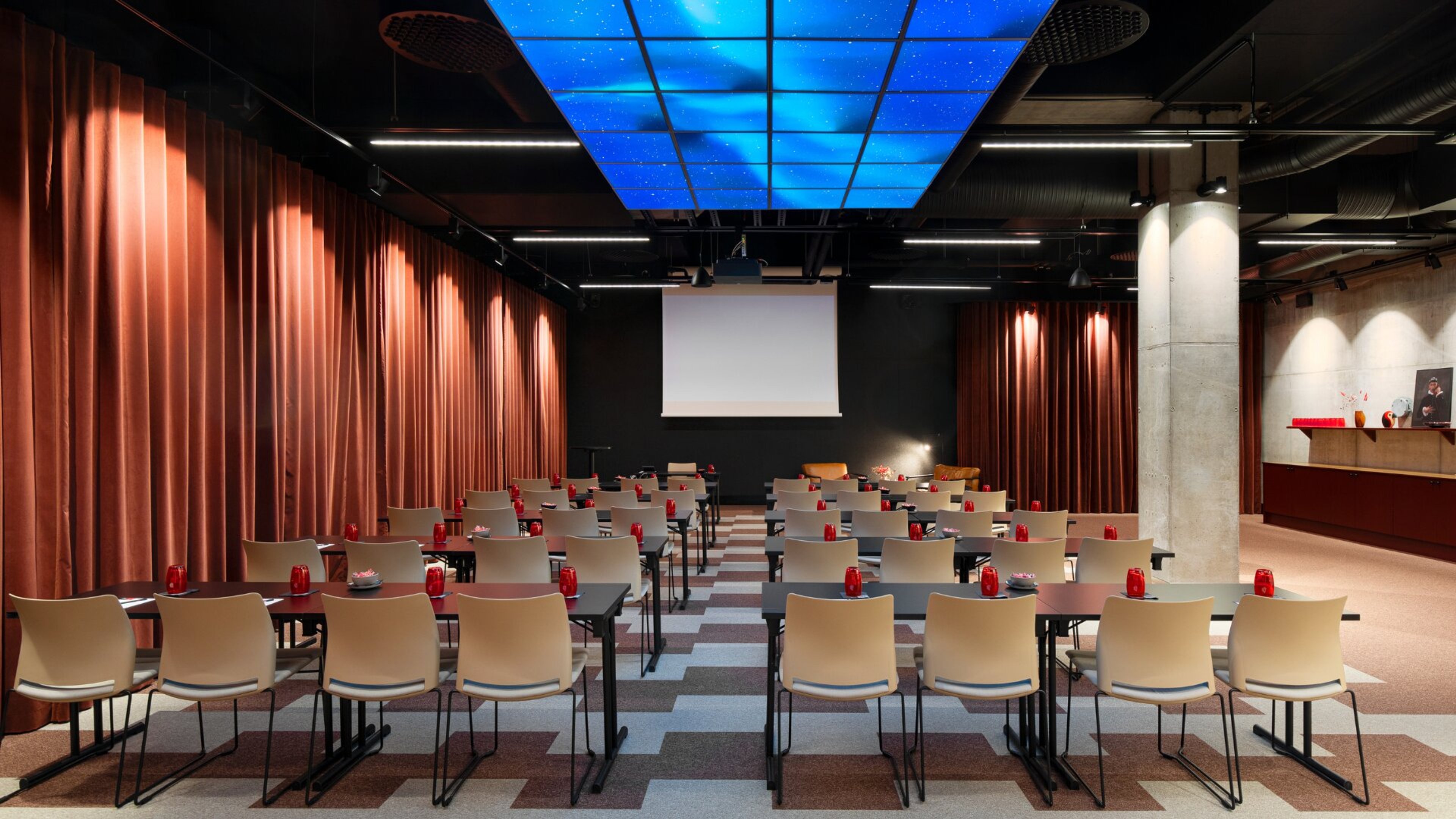 Moderni auditorio ruskeilla verhoilla ja sinisellä kattovalaistuksella. Valkoiset tuolit ja mustat pöydät rivistöissä, tasainen lattia ja projektorikangas edessä.
