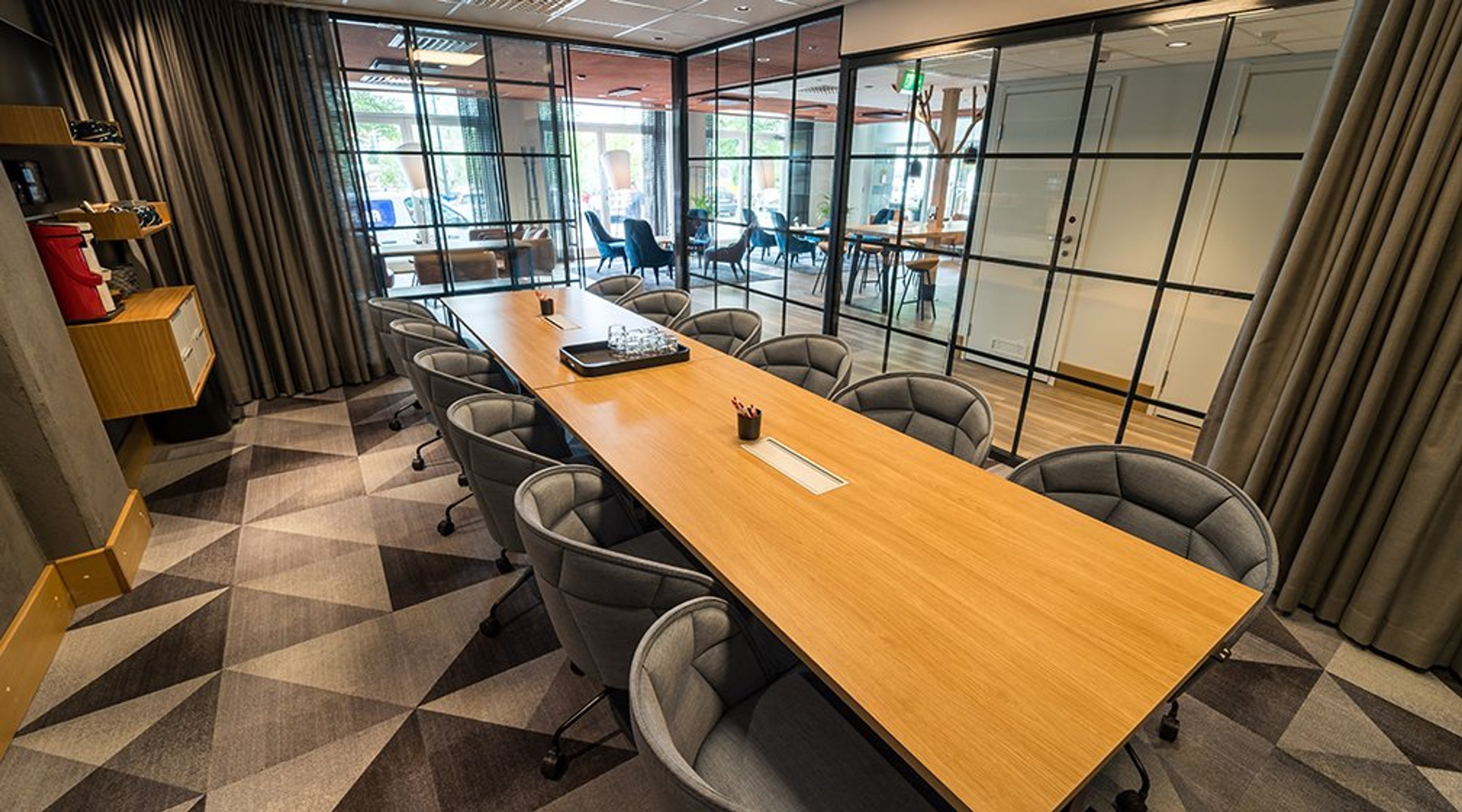 Moderni neuvotteluhuone, jossa puinen pöytä ja harmaat pyörälliset tuolit. Lasiseinän takana näkyy kahvilatila. Lattialla geometrinen kuviollinen matto.