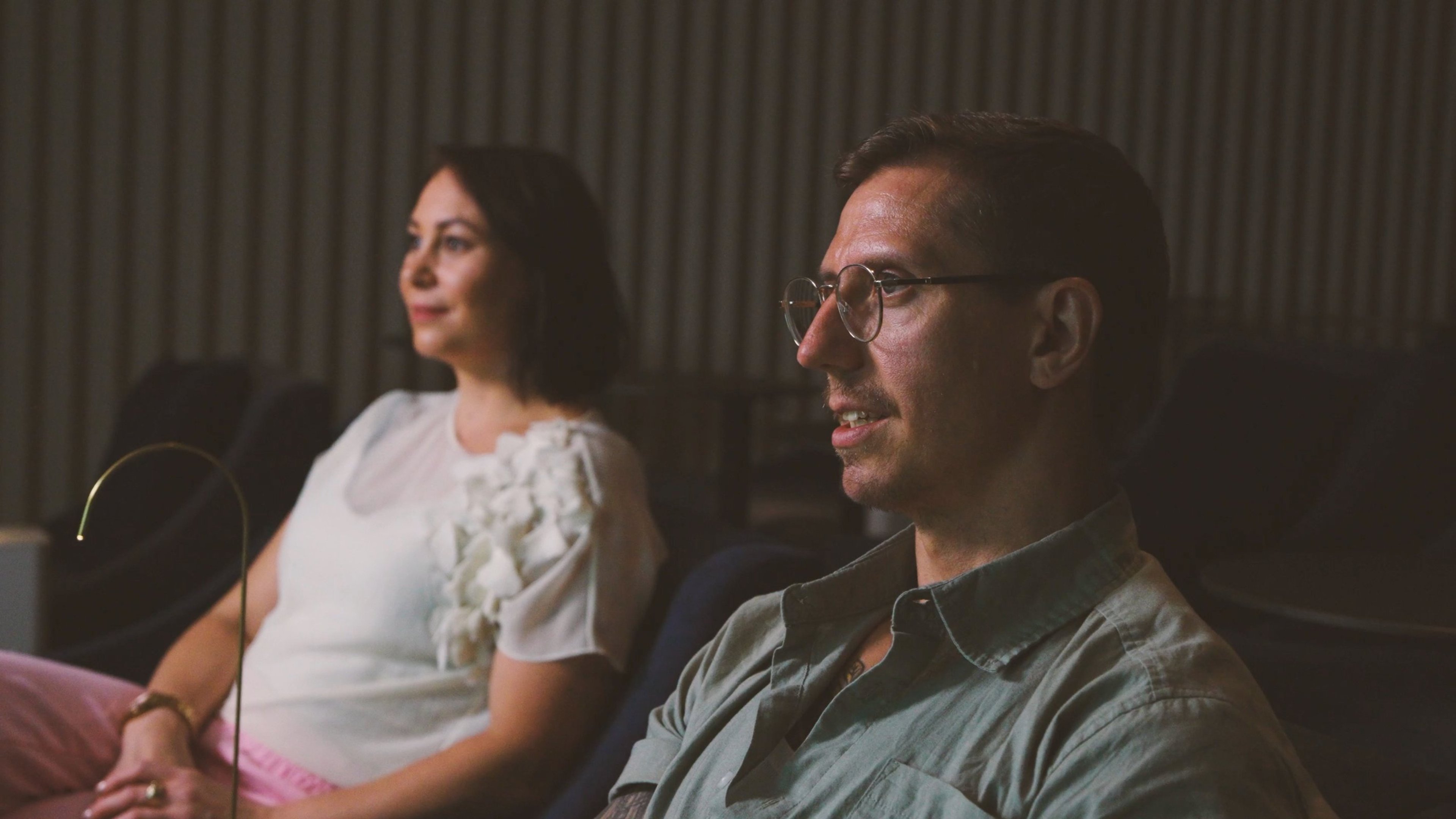 Kaksi henkilöä istuu auditoriossa hämärässä valaistuksessa, toinen valkoisessa paidassa ja toinen harmaassa kauluspaidassa silmälasit päässä.