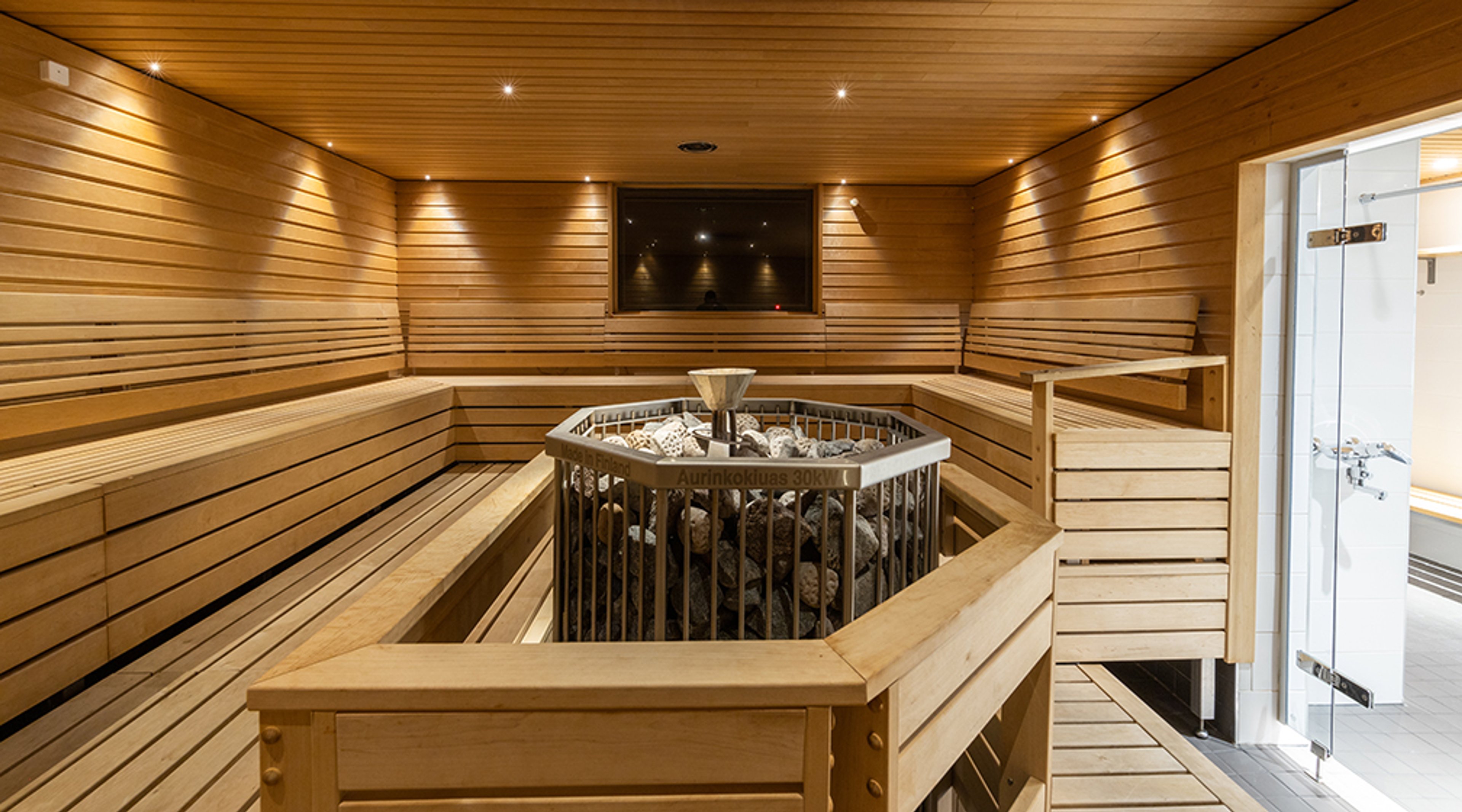 Moderni sauna puupaneloinnilla, keskellä kiuas kivikehyksen sisällä. Seinustoilla leveät lauteet ja katon rajassa pieniä valaisimia.