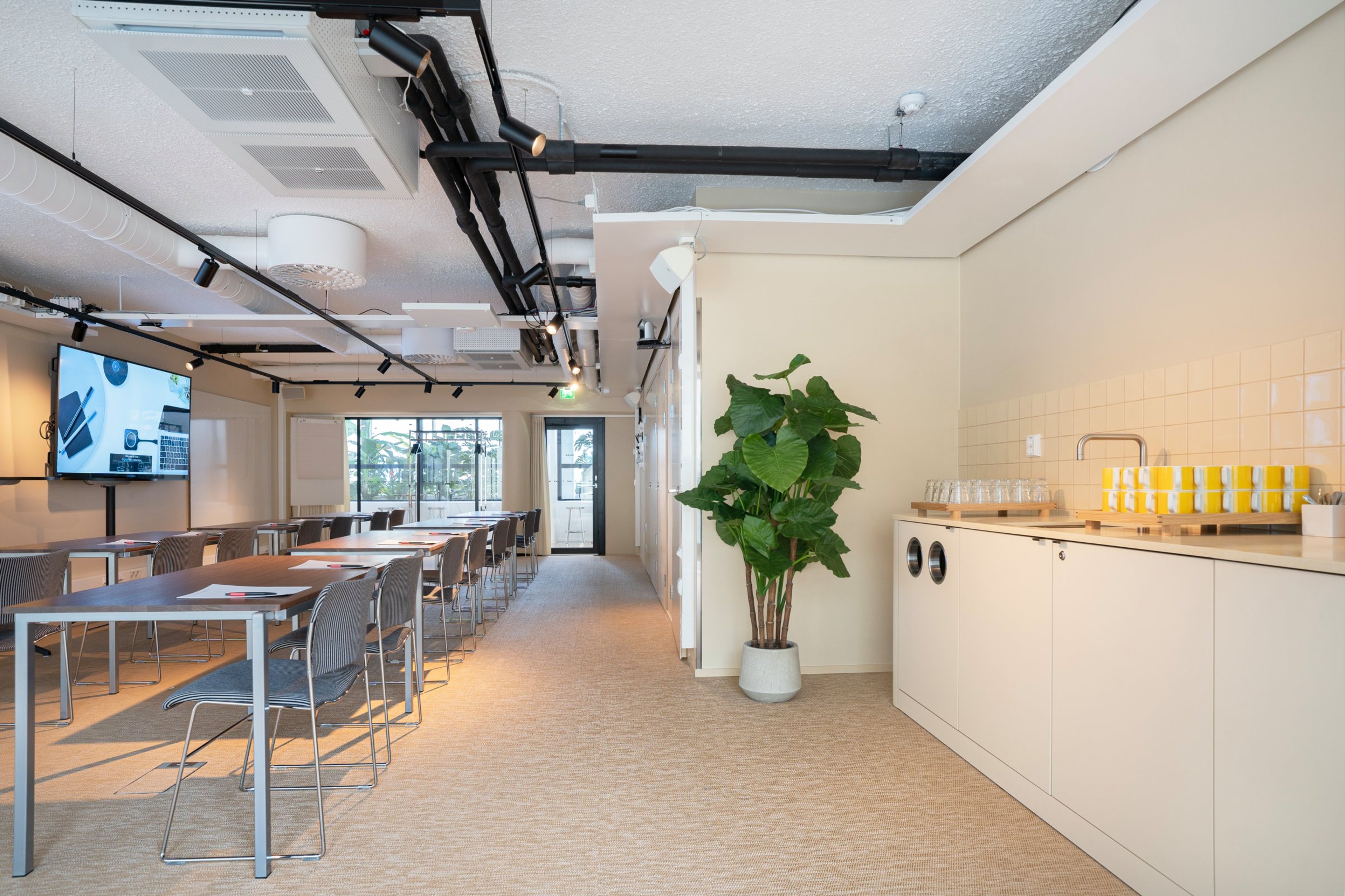 Moderni kokoustila, jossa pitkät pöydät, näyttö seinällä, isot ikkunat ja pieni keittiönurkkaus. Tasainen lattiapinta ja leveä käytävä.