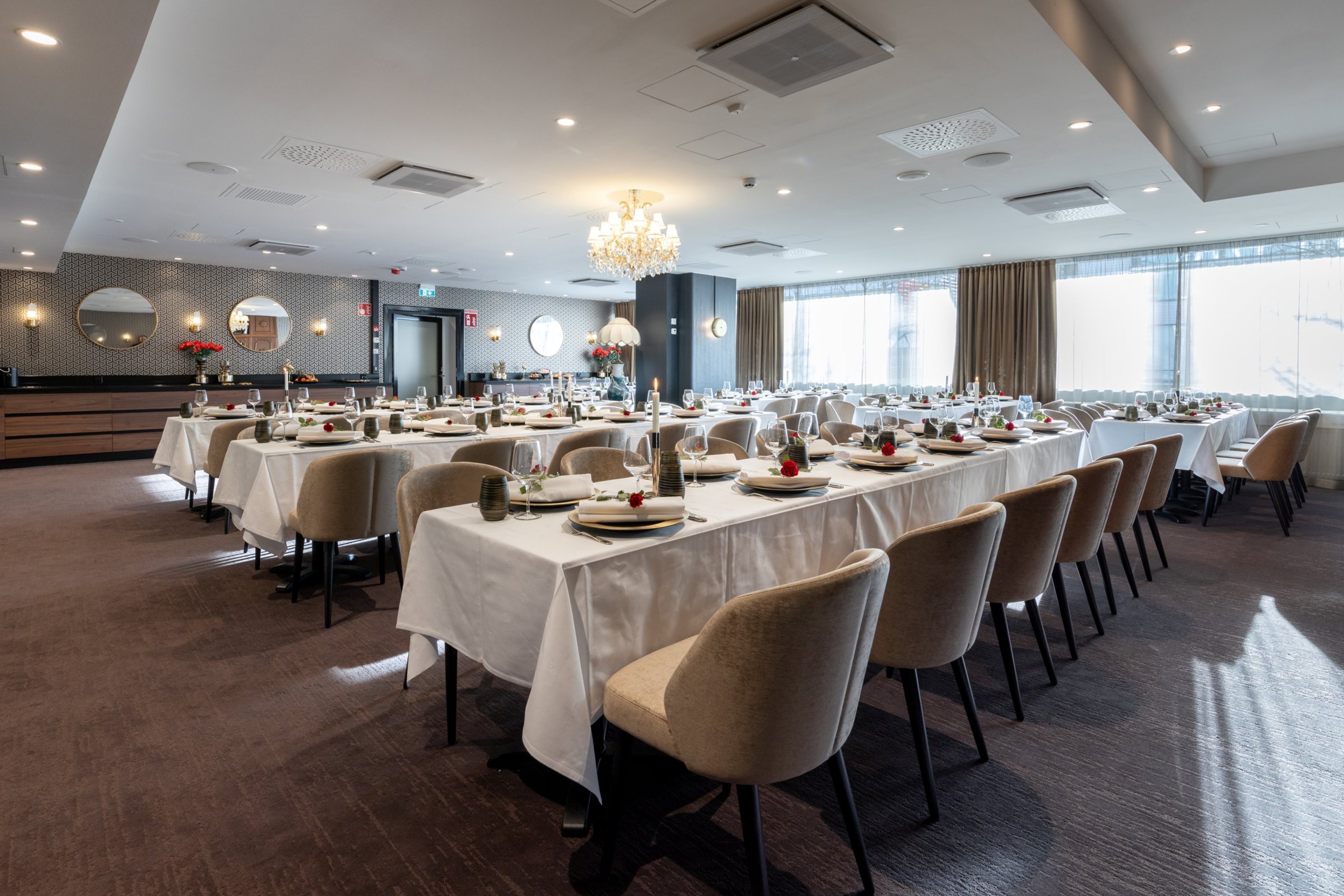 Tyylikäs juhlatila valkoisilla pöytäliinoilla ja vaalean beigeillä tuoleilla. Tilava sali on valaistu kattokruunulla ja isoilla ikkunoilla.