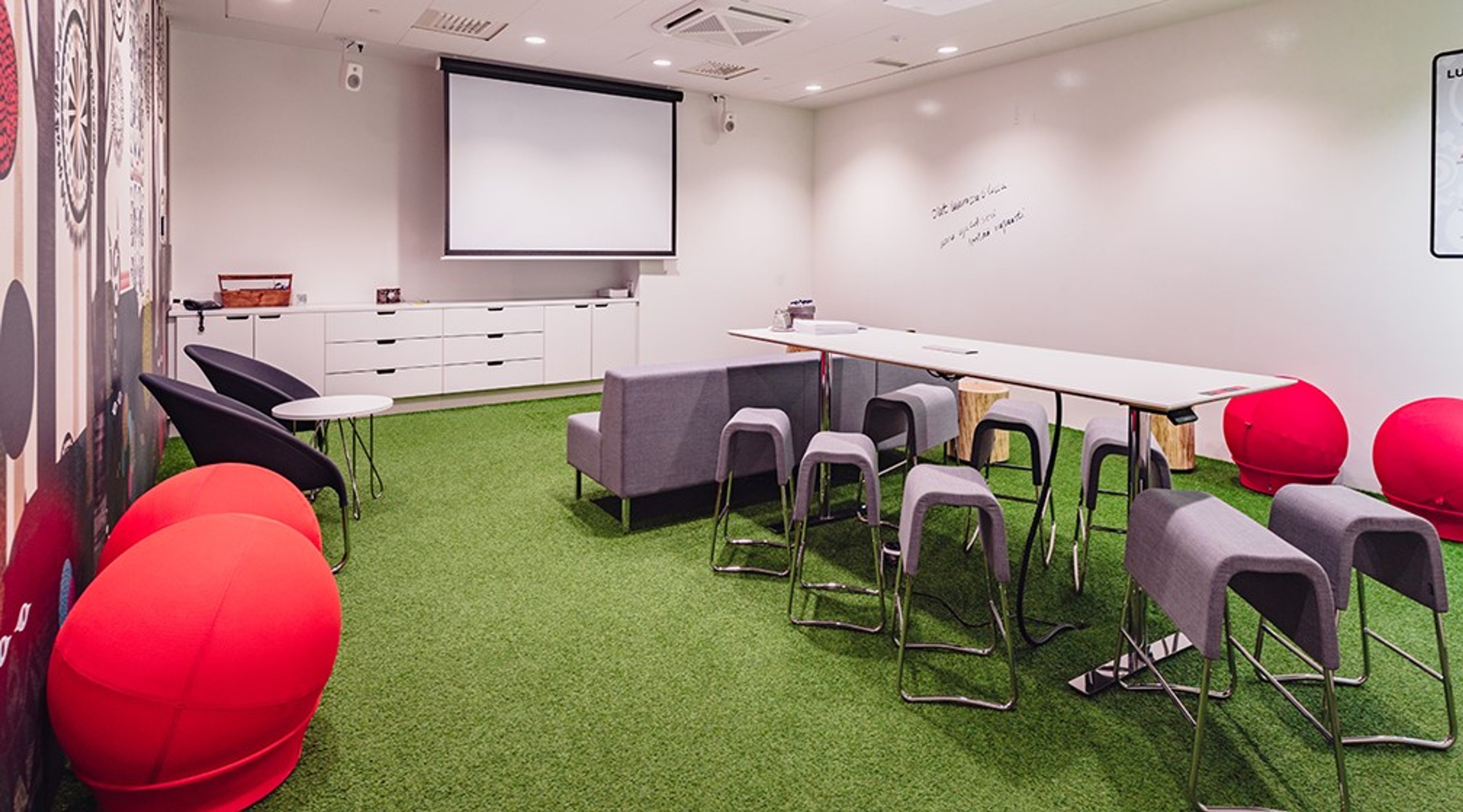Moderni kokoushuone vihreällä lattiamatolla, valkoinen pöytä, harmaita tuoleja ja punaisia istuinpalloja. Projektorikangas seinällä.