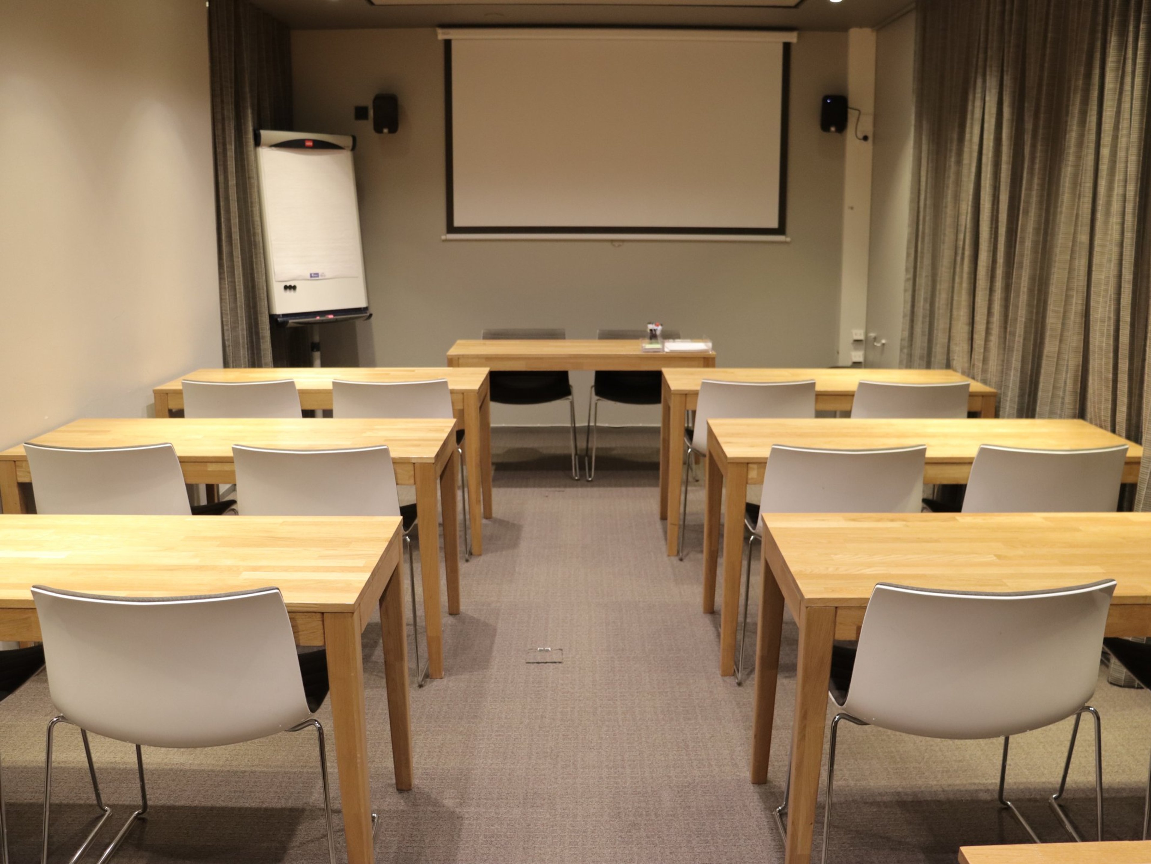 Pieni kokoushuone puisilla pöydillä ja valkoisilla tuoleilla, edessä valkokangas ja fläppitaulu. Huoneessa on tasainen kokolattiamatto.