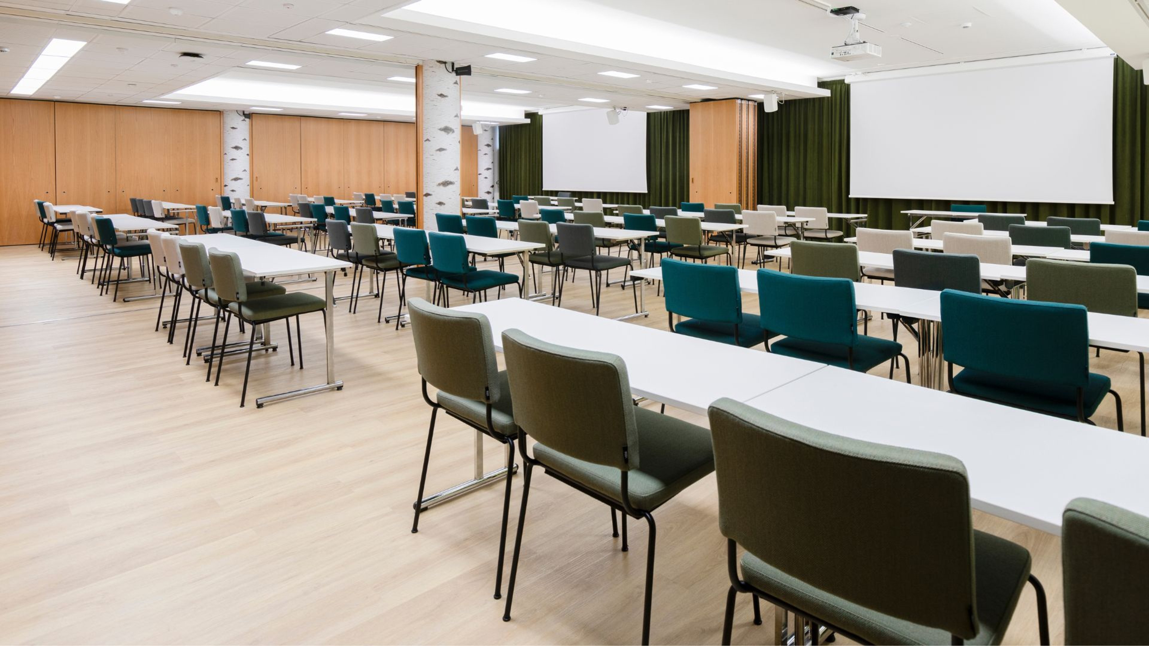 Valoisa kokoushuone, jossa rivit valkoisia pöytiä ja vihreitä sekä turkooseja tuoleja. Edessä kaksi valkokangasta ja puiset seinäpaneelit.