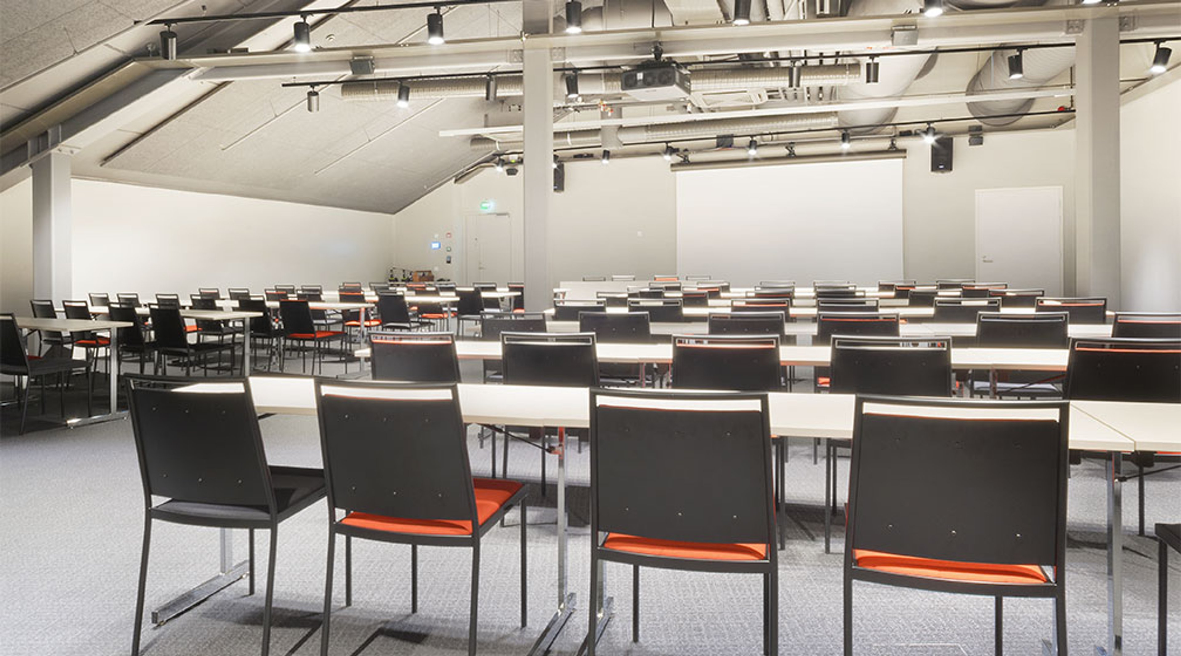 Suuri kokoustila pitkillä pöydillä ja mustilla tuoleilla, joissa punaiset istuimet. Valkoiset seinät ja hyvä valaistus katossa.