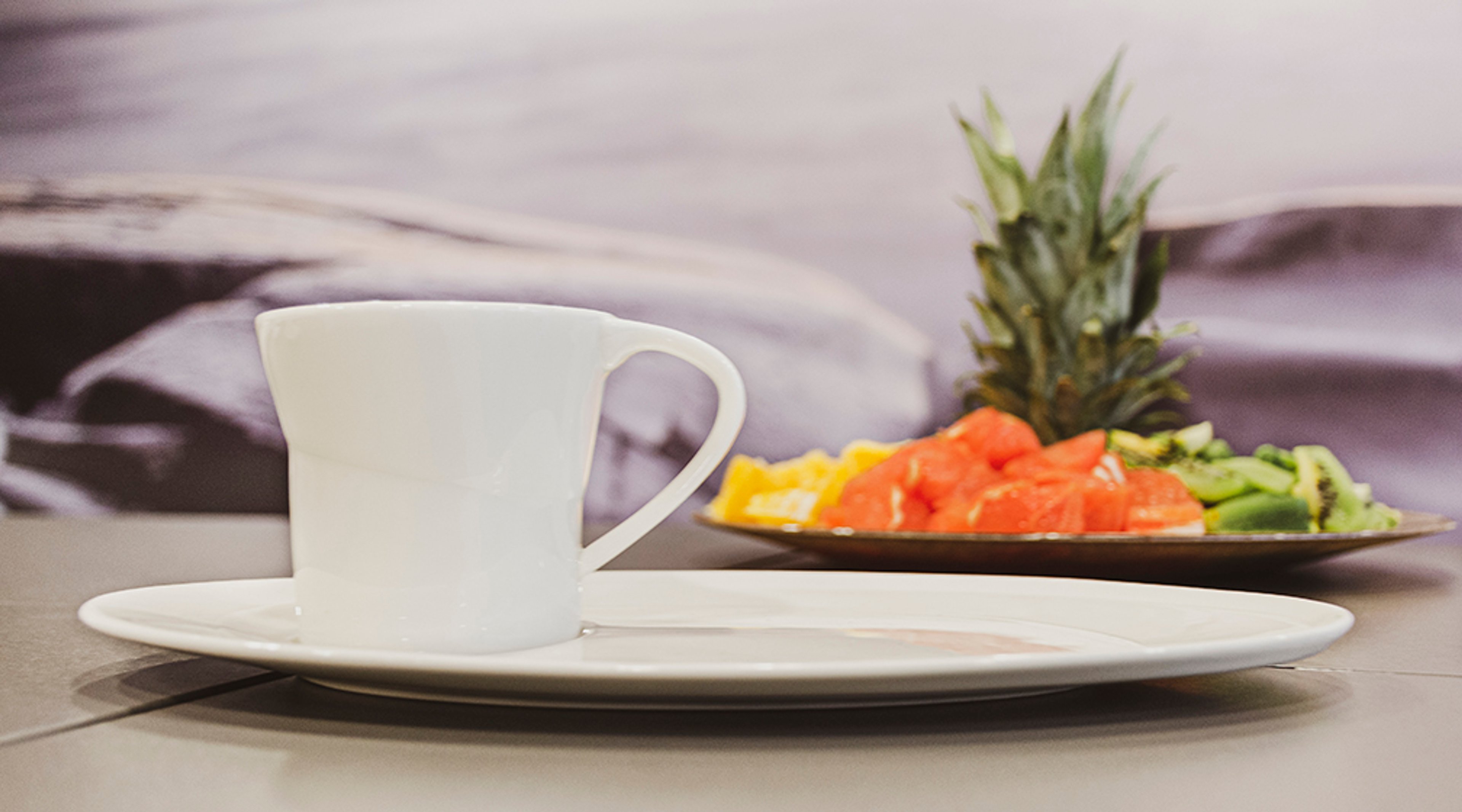 Valkoinen kahvikuppi lautasella, taustalla hedelmälautanen, jossa vesimelonia, ananasta ja kiiviä.