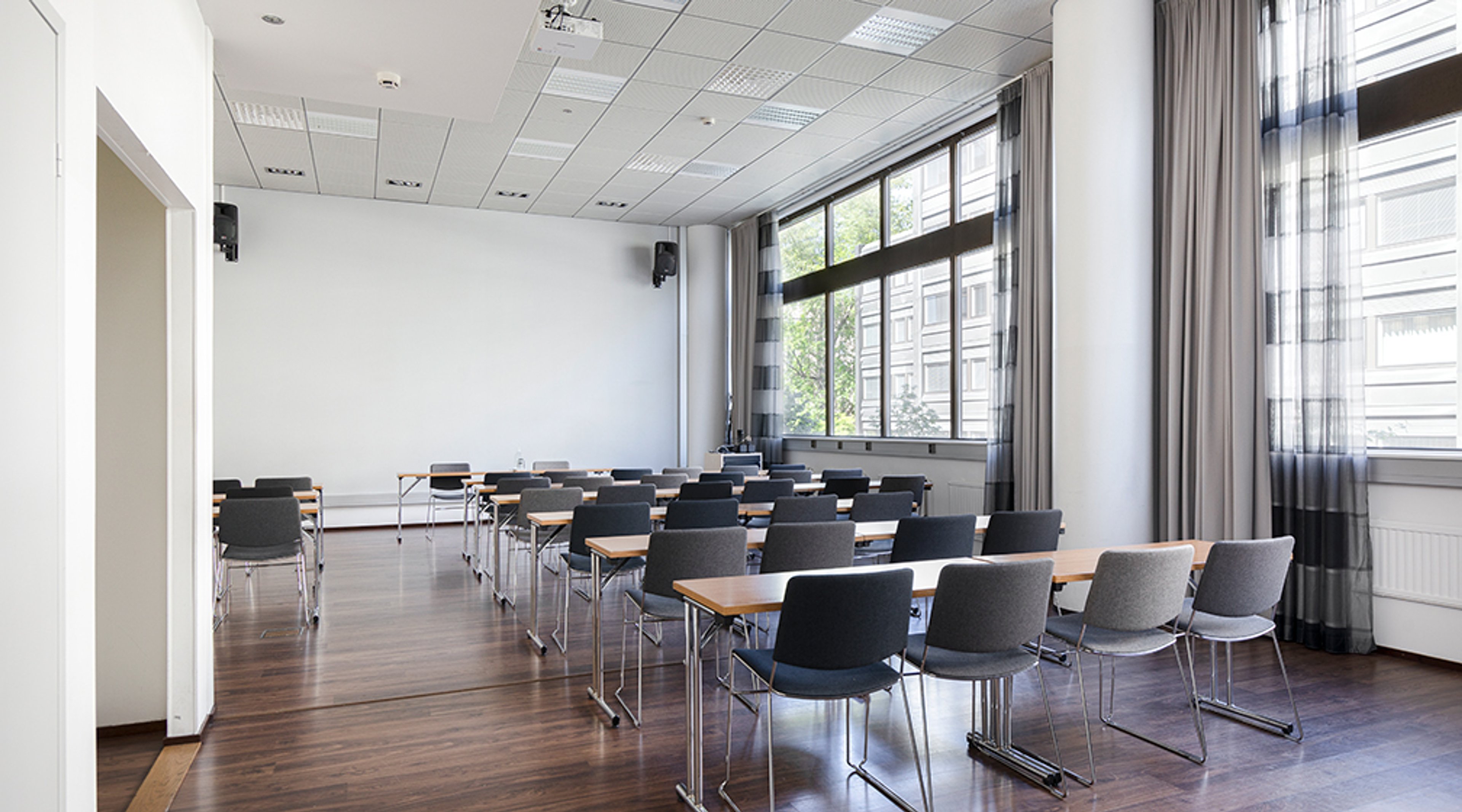 Moderni kokoushuone, jossa pitkät pöydät ja tuolit riveissä, isot ikkunat, puulattia ja vaaleanharmaat verhot.