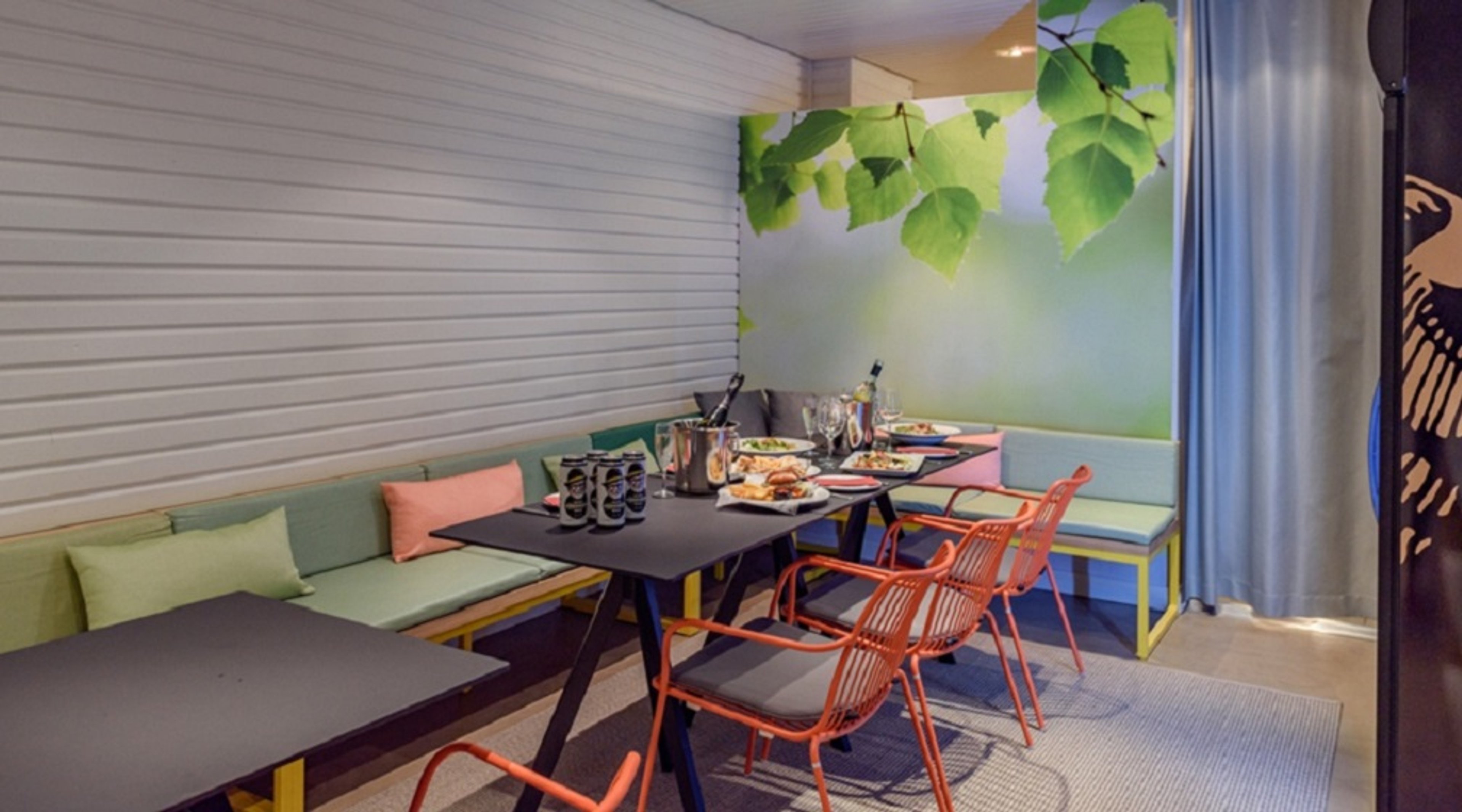 Moderni ruokailutila, jossa vihreät sohvat, oranssit tuolit ja musta pöytä. Seinällä luontoaiheinen lehtikuvioinen tapetti.