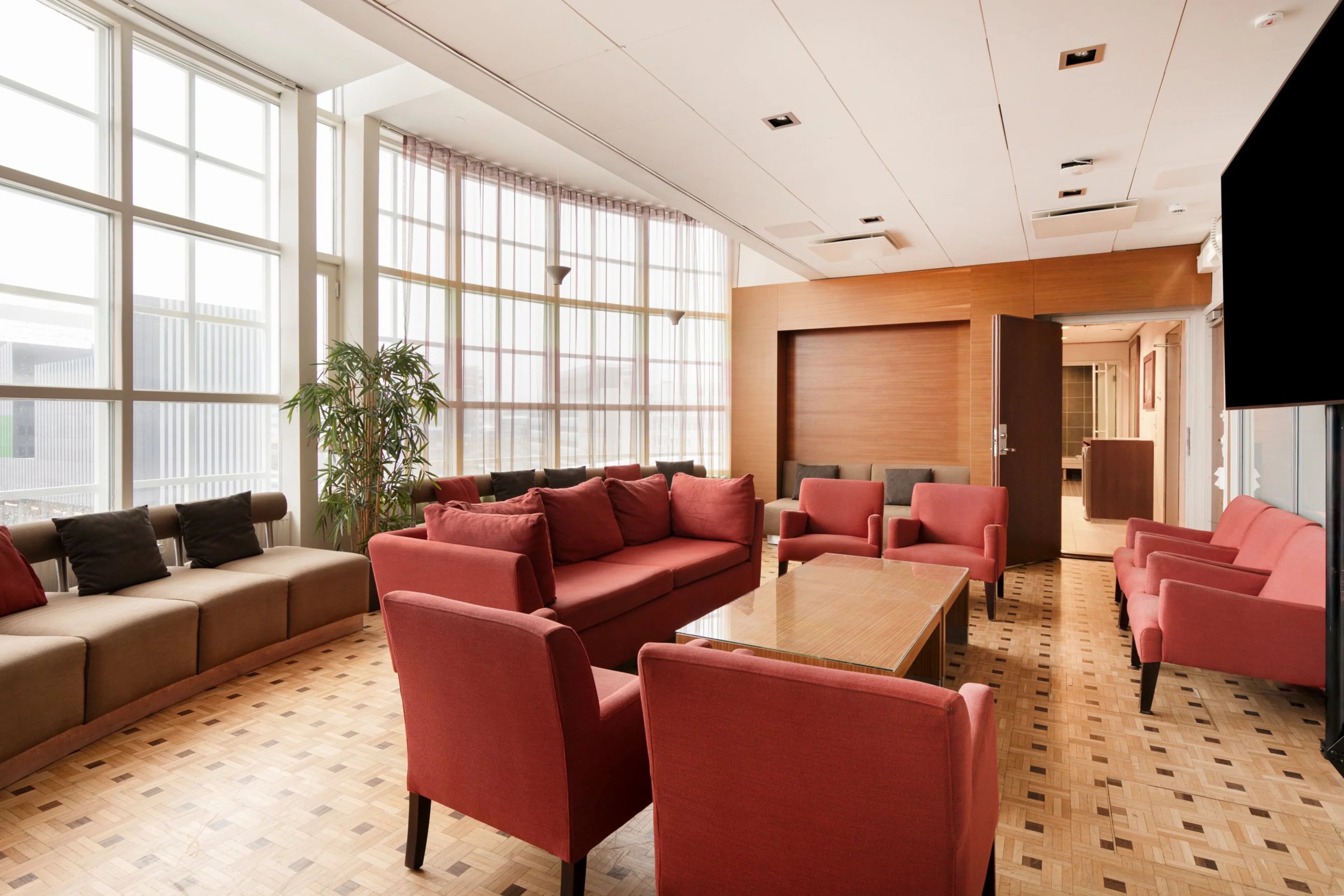 Valoisa lounge-tila, jossa punaisia nojatuoleja ja sohvia, ruskea sohva, puinen pöytä ja puulattia. Suuret ikkunat ja korkea katto.