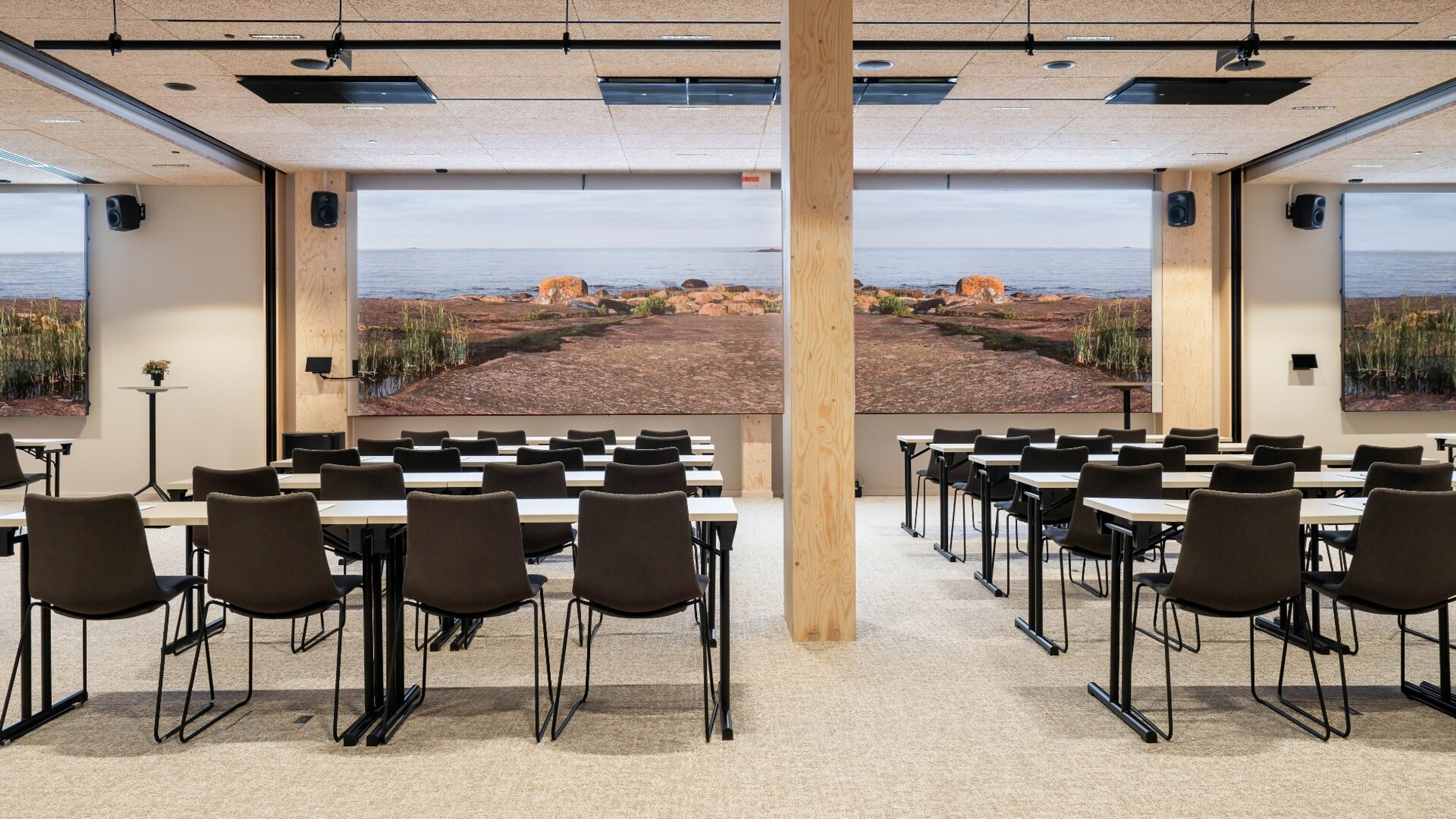 Moderni luentosali, jossa pöydät ja tuolit riveittäin. Takaseinällä suuri näyttö merellisellä maisemalla. Lattia on tasainen ja tilassa puinen tukipylväs keskellä.