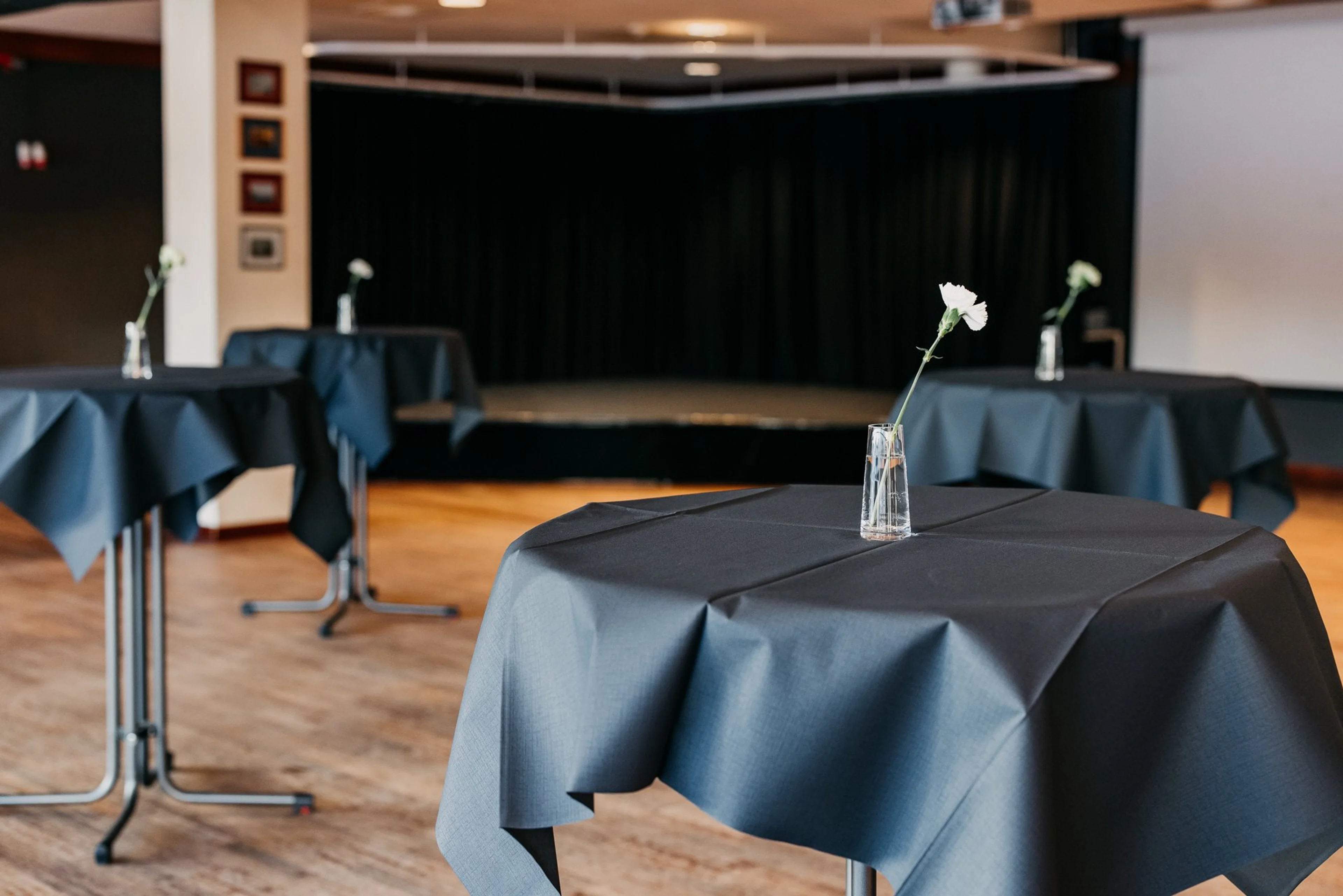 Tapahtumapaikka pöytiä tummilla liinoilla ja maljakoilla, joissa on valkoisia kukkia. Taustalla näyttämö tummalla verholla.