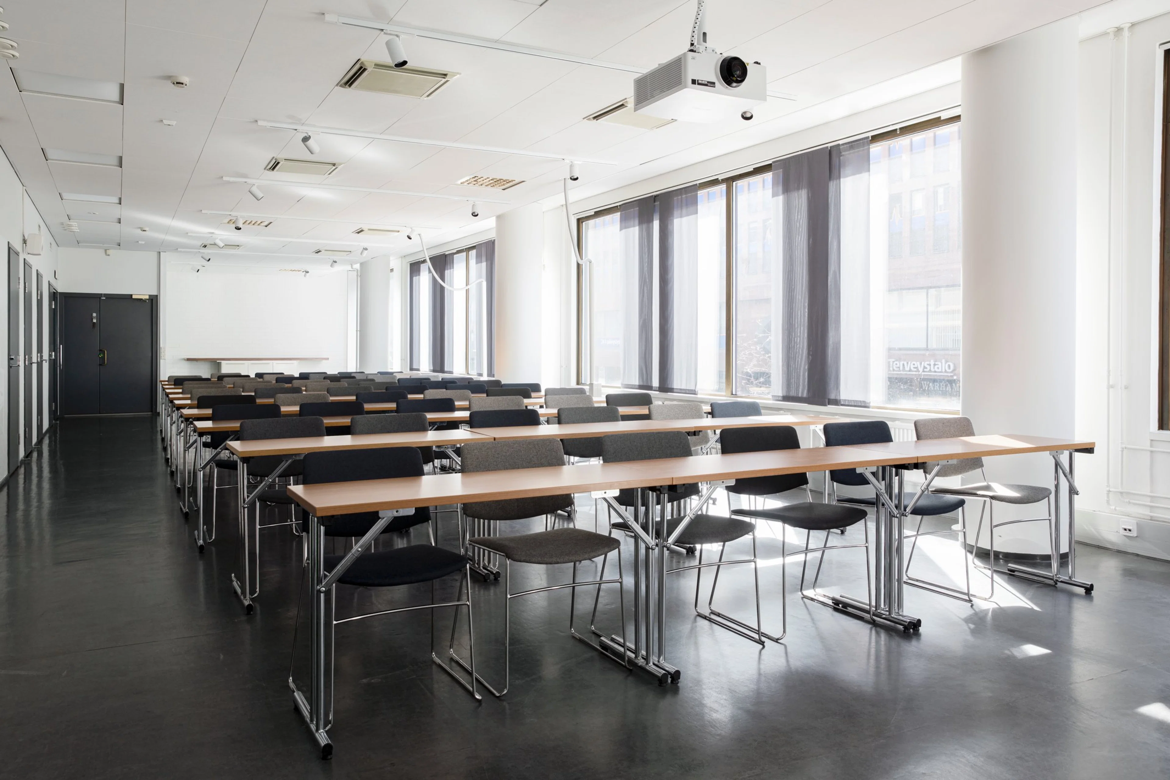 Nykyaikainen kokoustila riveittäin järjestetyillä pöydillä ja tuoleilla. Vaalea huone, jossa isot ikkunat ja kiinteä projektori katossa. Tasainen lattia.