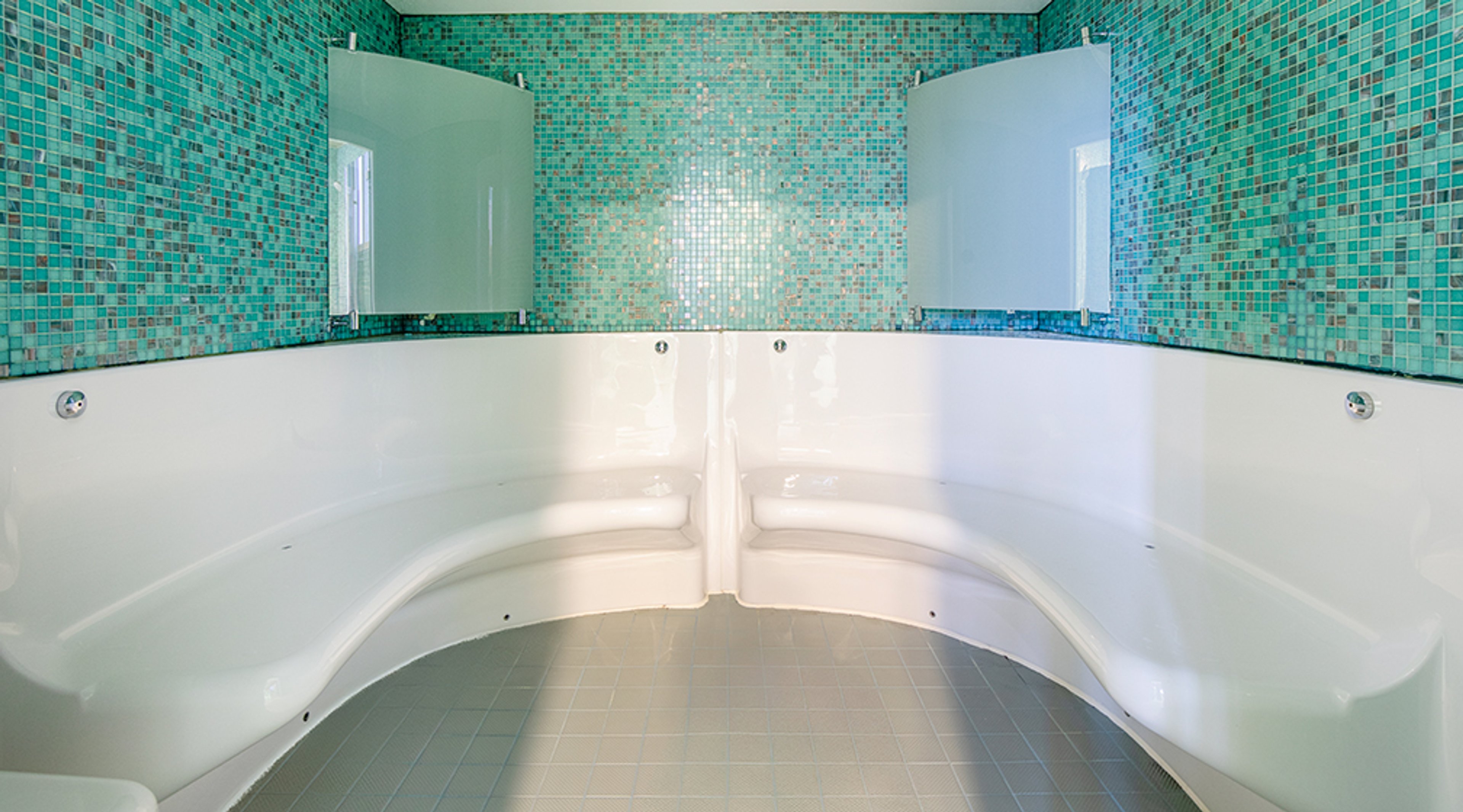 Turkoosin mosaiikkiseinän eteen asetettu valkoinen kylpyallas, jossa on kaksi erillistä istumapaikkaa ja läpinäkyvät suojalevyt.