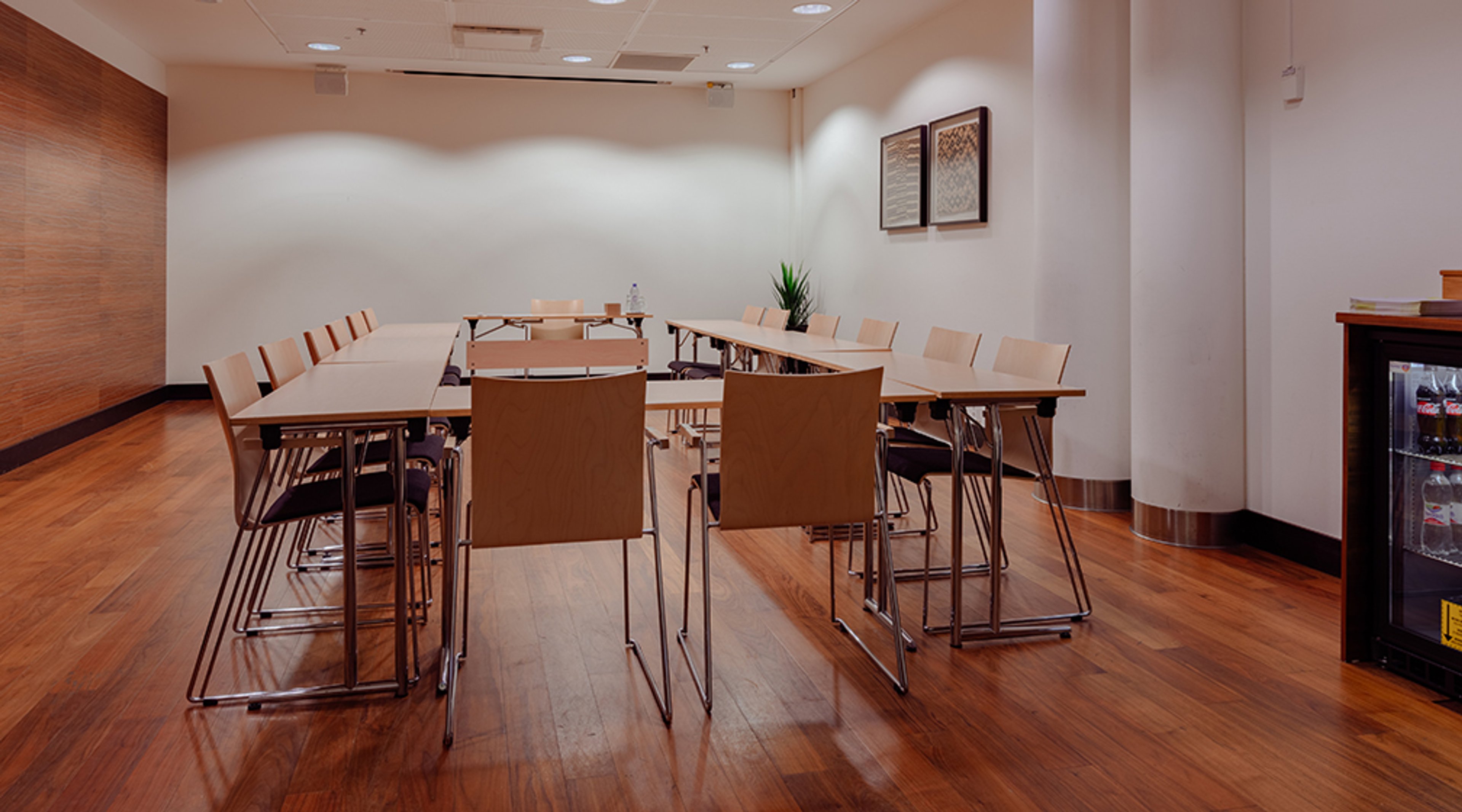 Neuvotteluhuone, jossa U-muotoinen pöytäjärjestely ja vaalea sisustus. Puulattia, seinällä taulut ja nurkassa pieni jääkaappi.