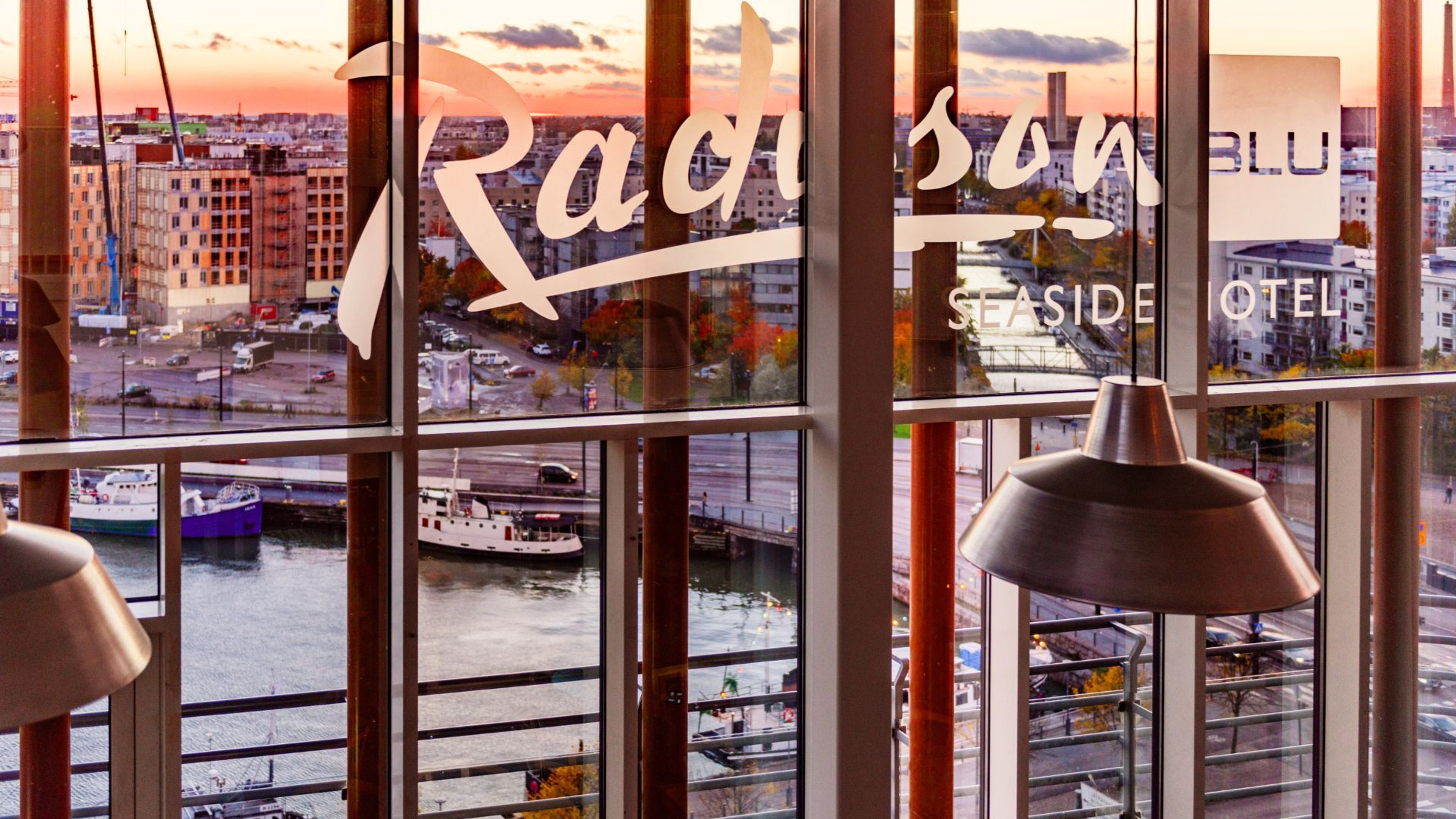Näkymä hotellin ikkunasta satamaan auringonlaskun aikaan. Etualalla modernit riippuvalaisimet, taustalla veneitä laiturin vieressä.