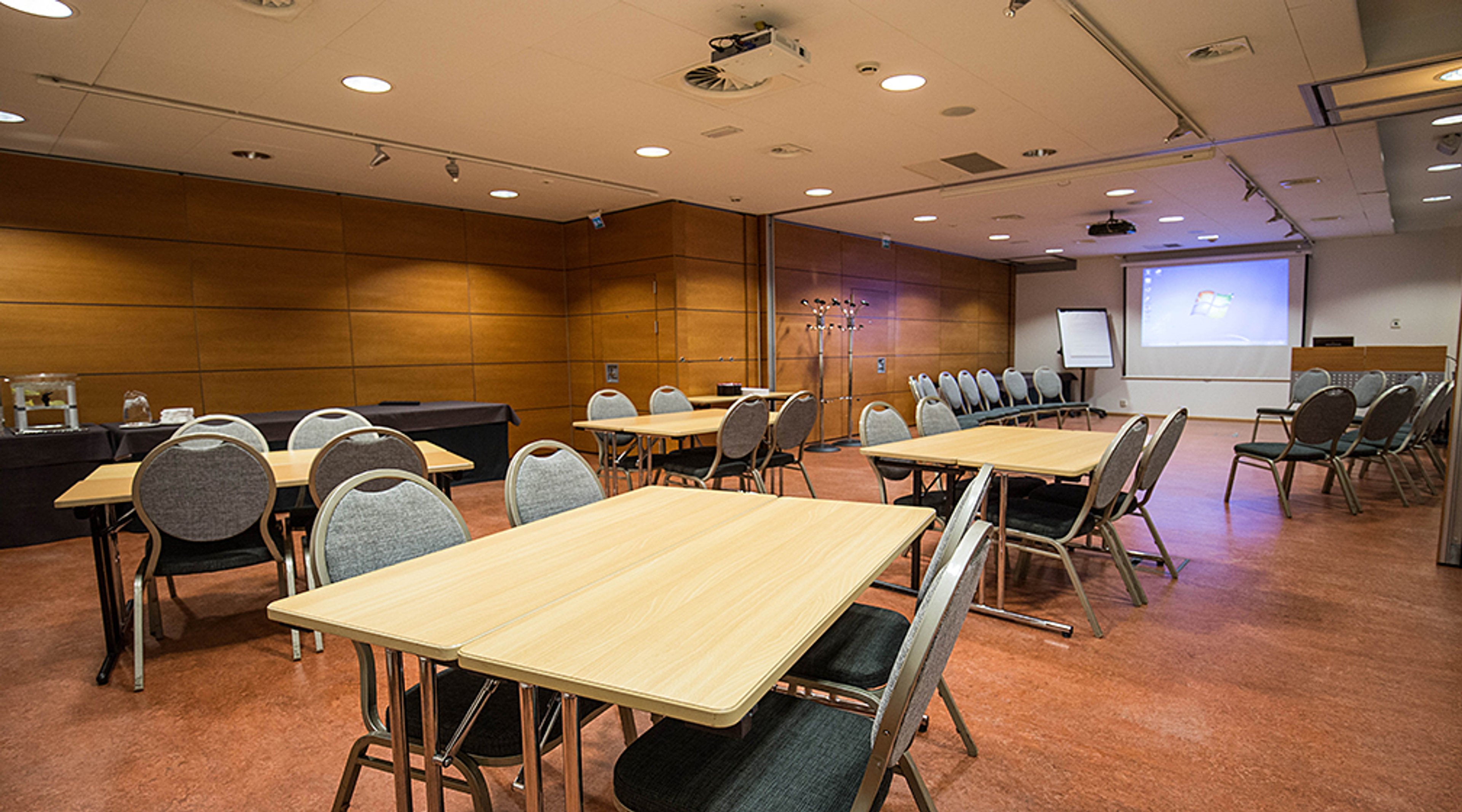 Kokoustila, jossa puiset pöydät, harmaat tuolit, puupaneelit seinillä, projektori ja valkokangas. Lattialla tasainen, punaruskea pinta.