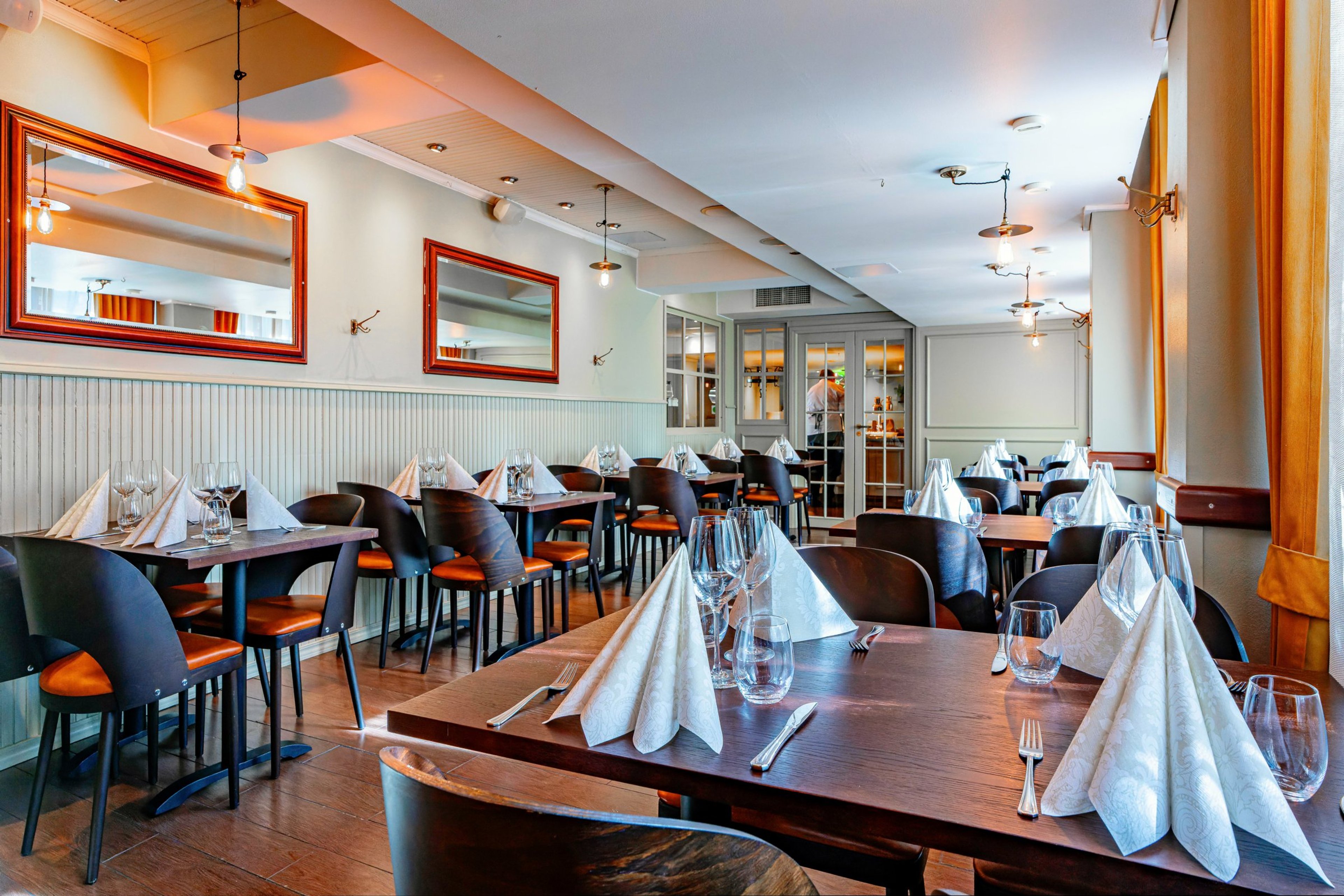 Ravintolan sisätila, jossa puiset pöydät on katettu valmiiksi. Tummat tuolit ja oranssi verho. Tila on valoisa puulattialla.