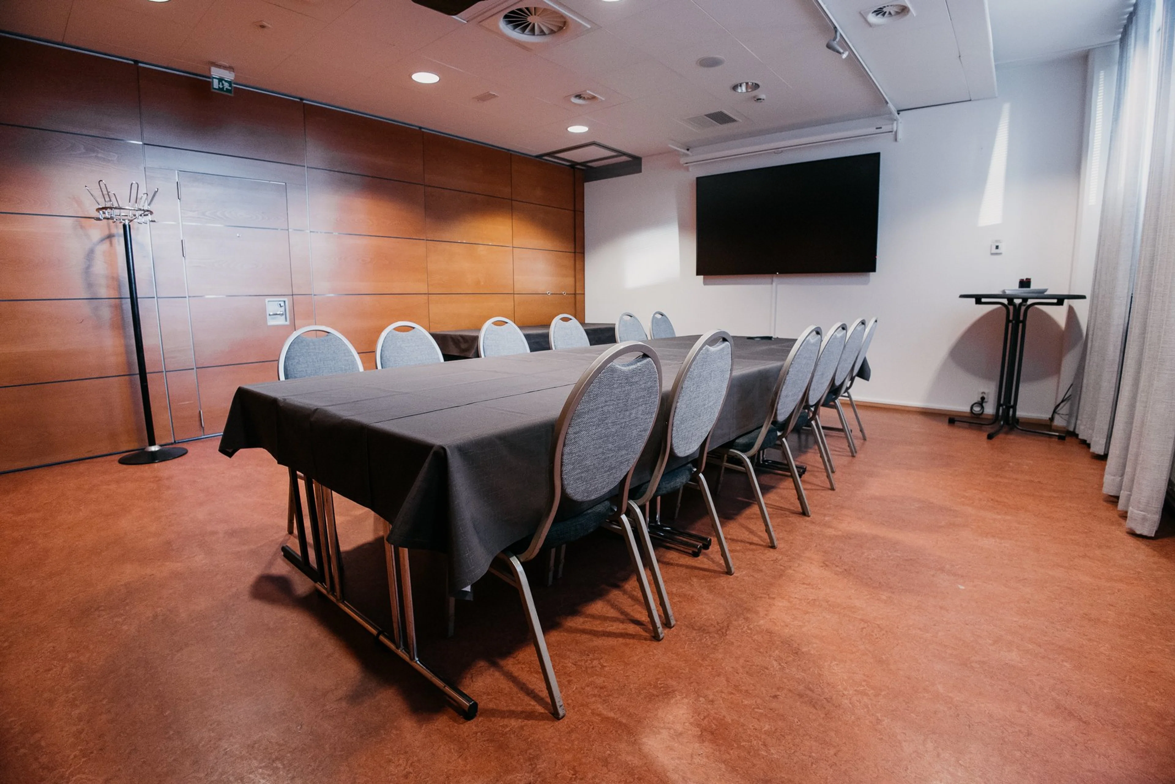 Kokoushuone puisilla seinäpaneeleilla, pitkä pöytä mustalla liinalla ja harmaat tuolit. Seinällä näyttö, lattia tasainen ja kova.