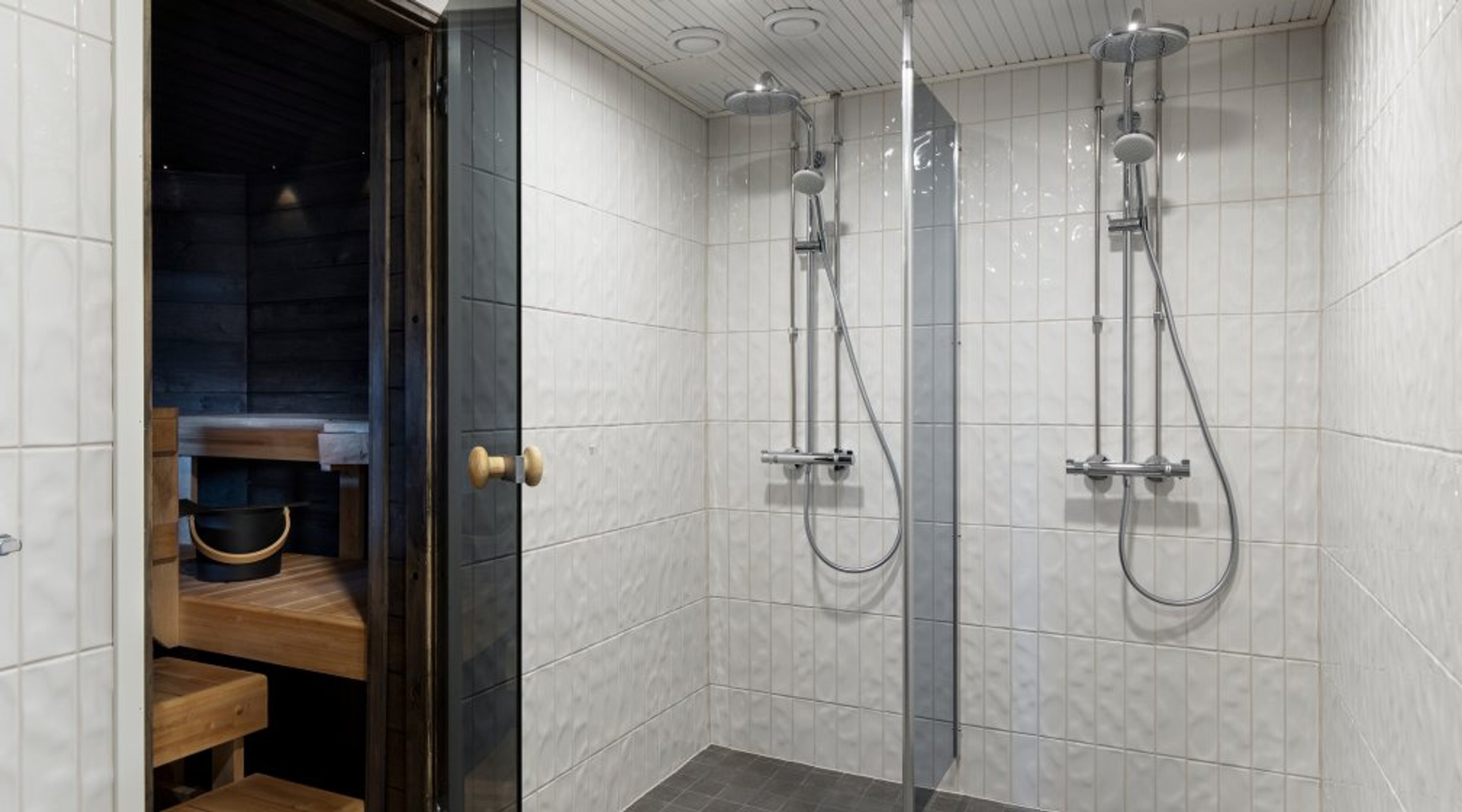 Light-tiled shower room with two showers. Door opens to adjacent sauna with wooden benches.