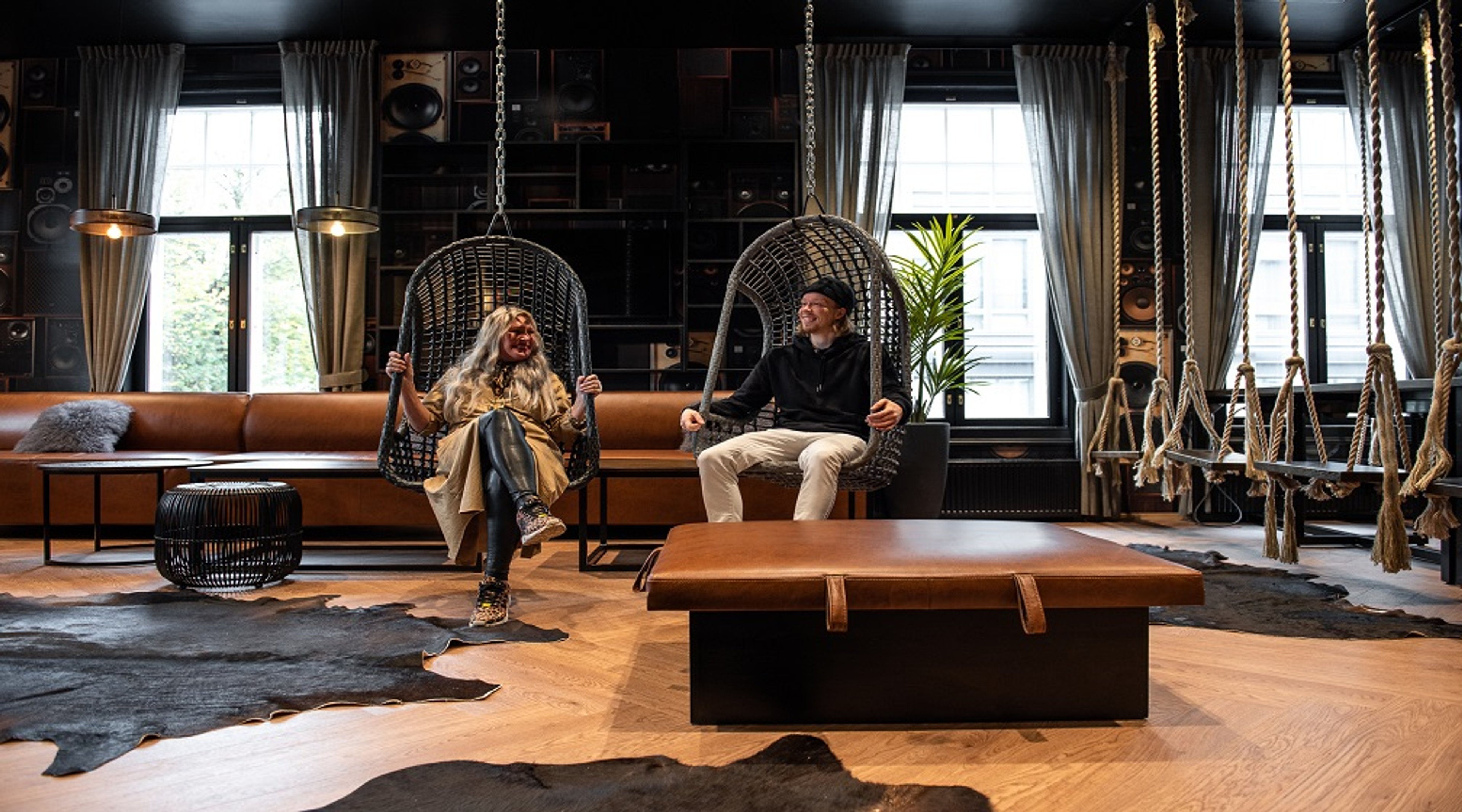 Tyylikkäästi sisustettu tila, jossa kaksi henkilöä istuu riippuvissa korihuonekaluissa. Huoneessa on nahkasohva, parkettilattia ja suuret ikkunat.