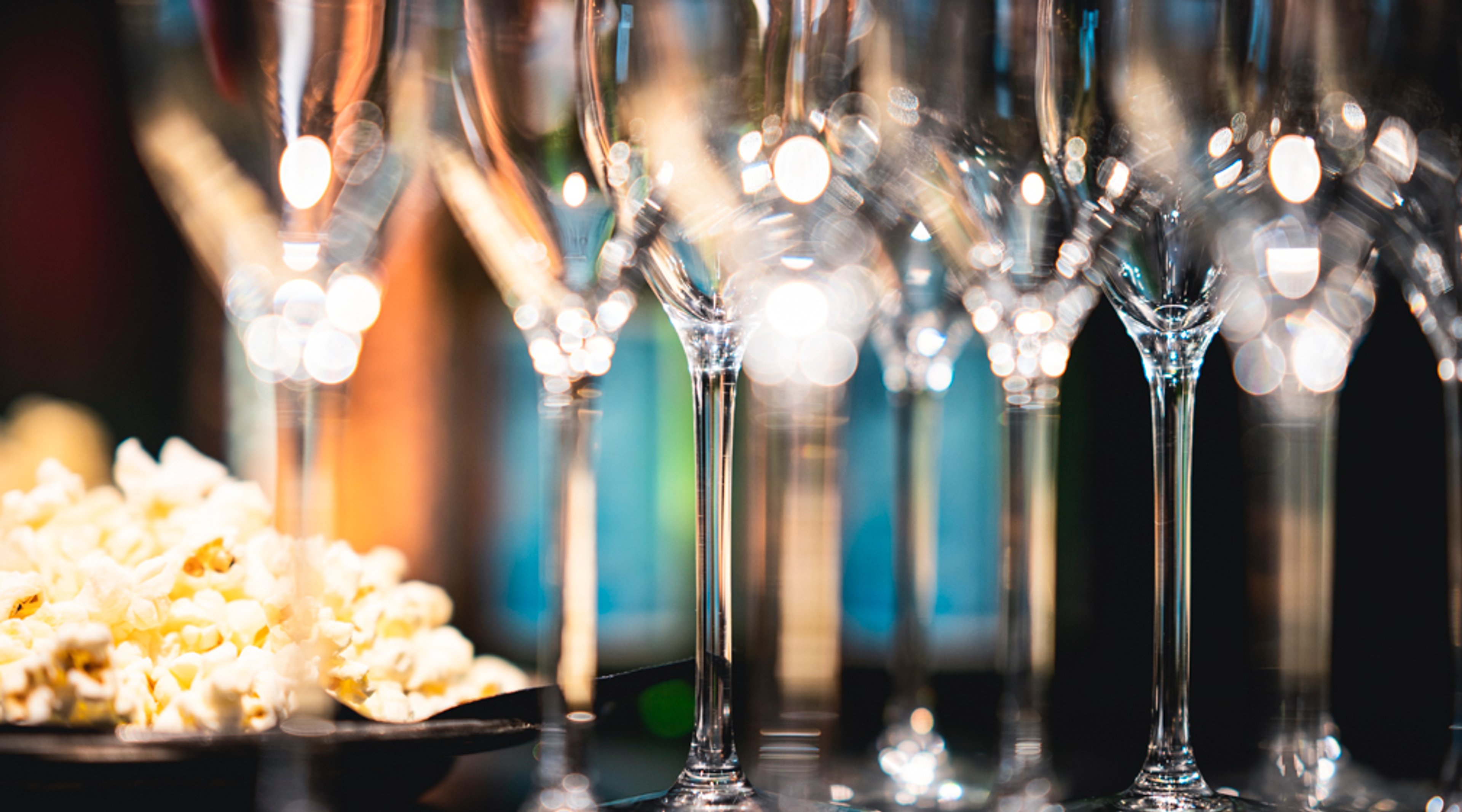 Rivistö tyhjiä samppanjalaseja, taustalla bokeh-valoefektejä, vasemmassa reunassa lautasellinen popcornia.