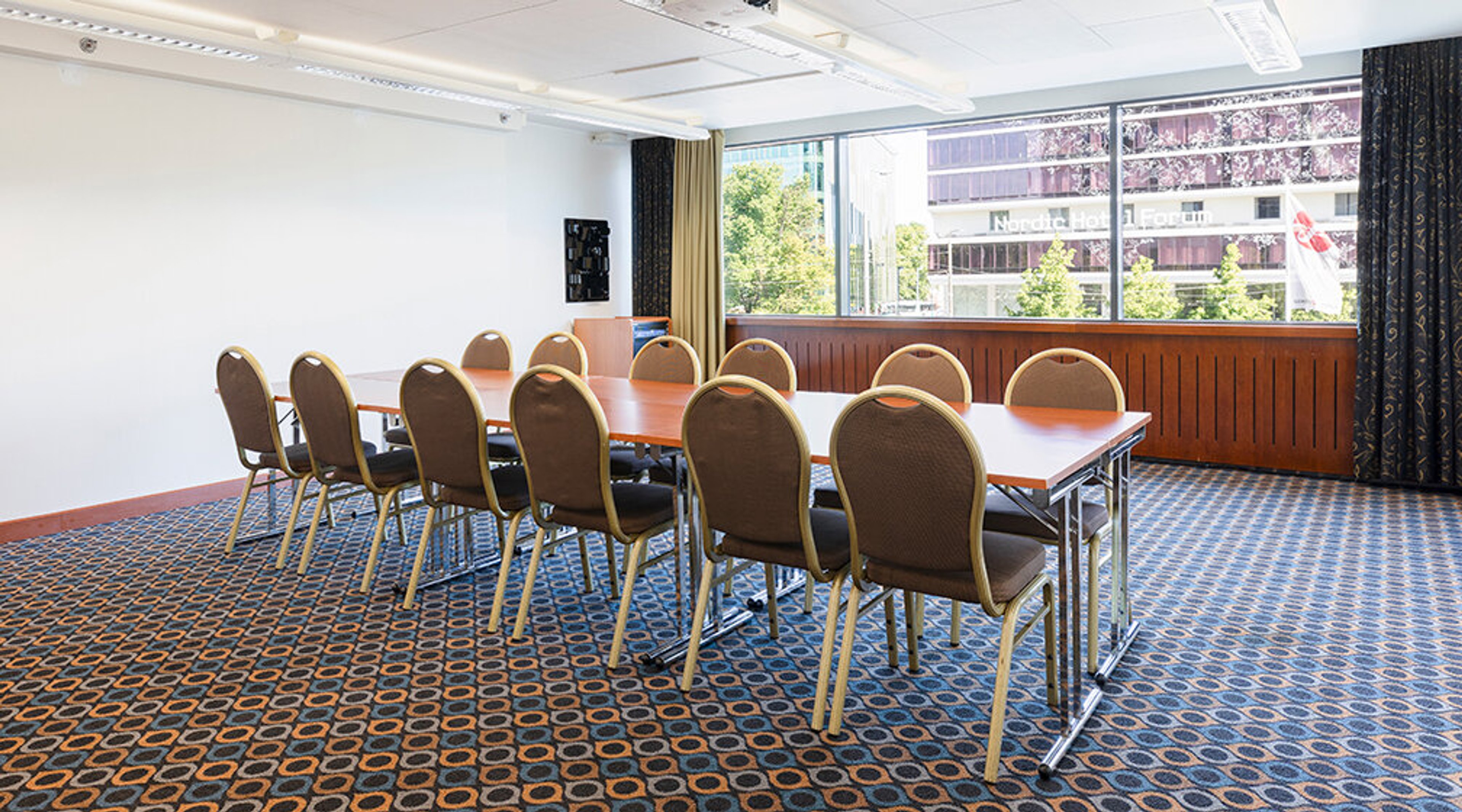 Conference room with long table and chairs, patterned wall-to-wall carpet, large window and even lighting.