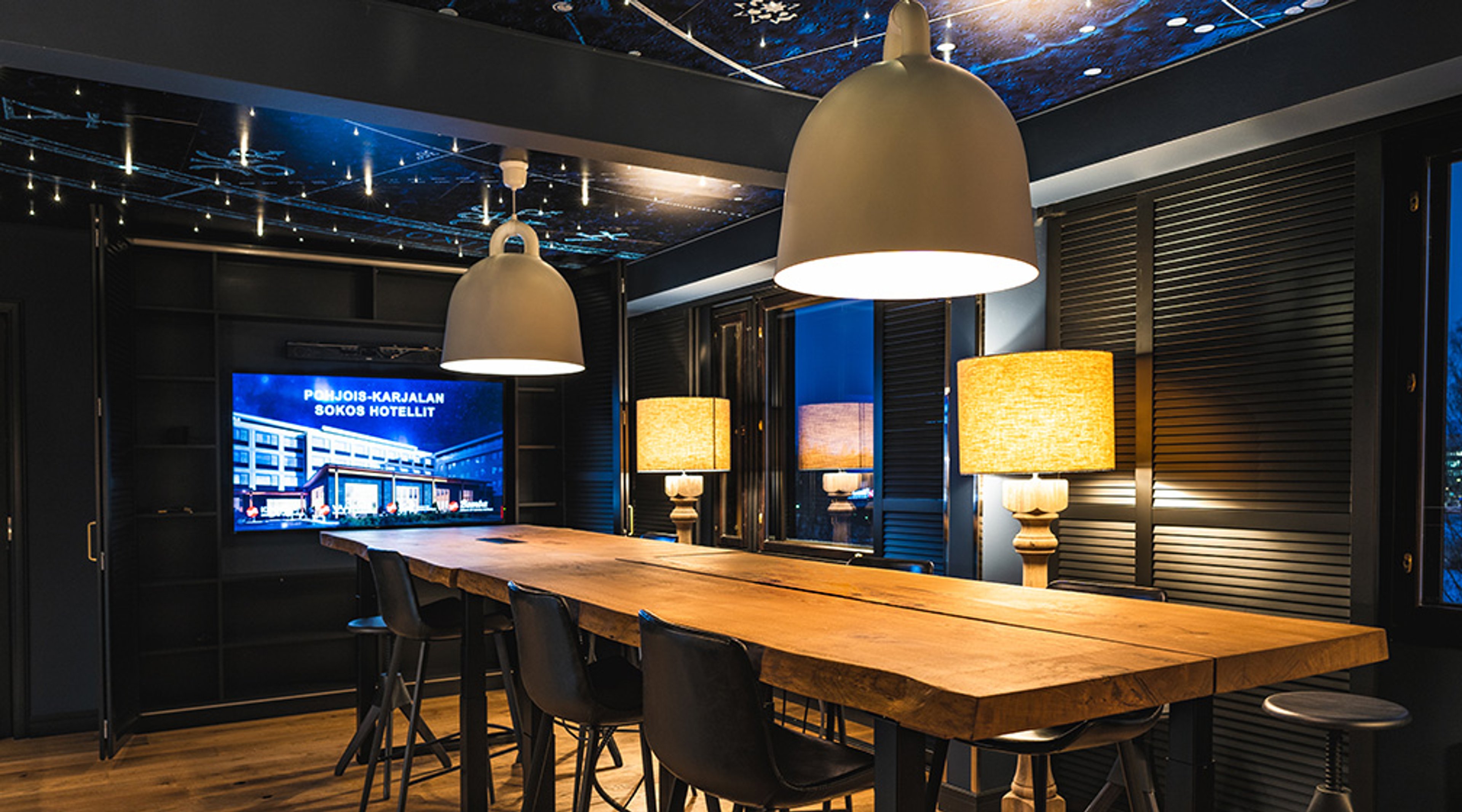 Tyylikäs kokoustila, jossa puinen pöytä, mustat tuolit, kaksi riippuvaa valaisinta ja näyttö takaseinällä. Kauniisti valaistu sininen katto.
