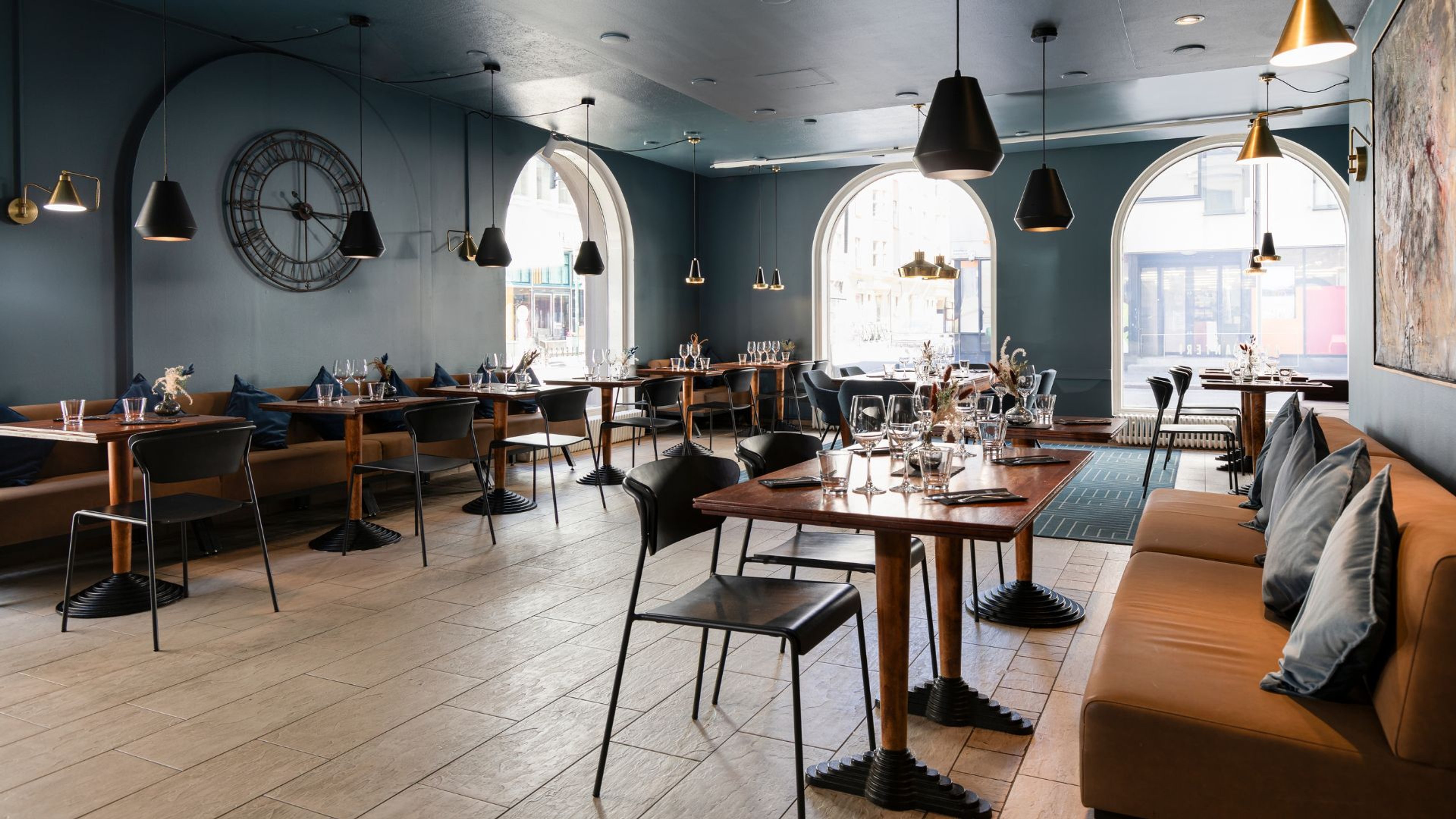 Tyylikkäästi sisustettu ravintola sinivihreillä seinillä, puulattialla ja suurilla kaartuneilla ikkunoilla. Puiset pöydät ja mustat tuolit.