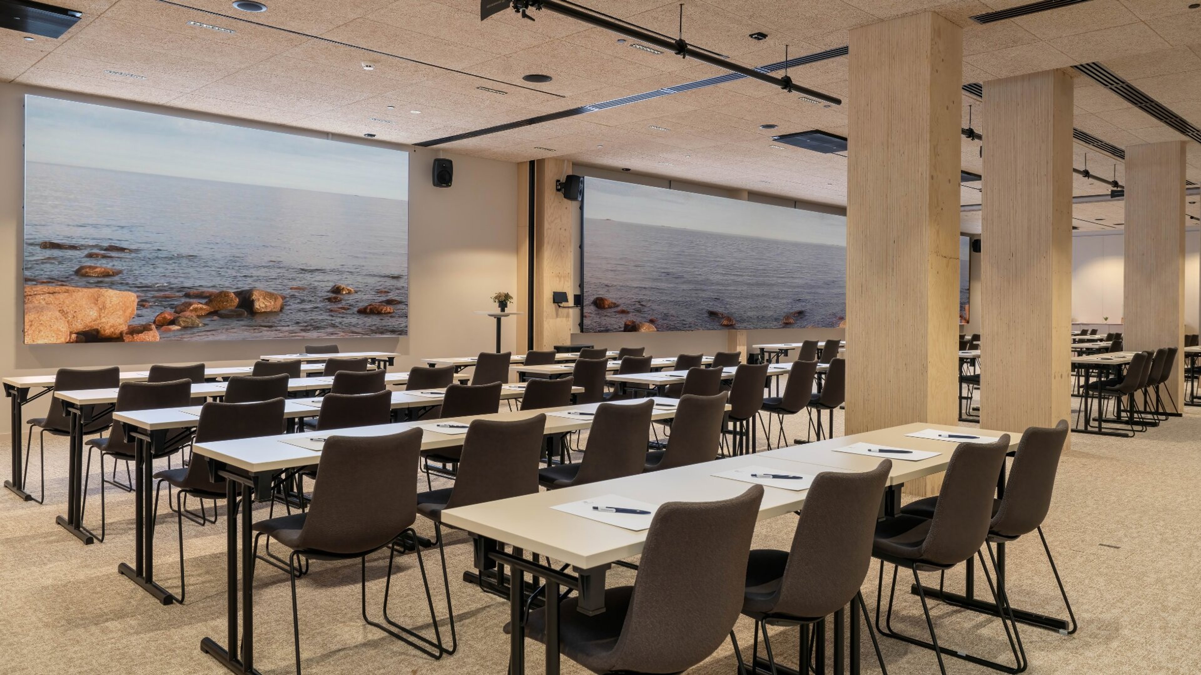 Moderni kokoustila, jossa pöytiä ja tuoleja. Seinällä suuri merimaisema. Tilassa puupilarit ja tasainen lattia.