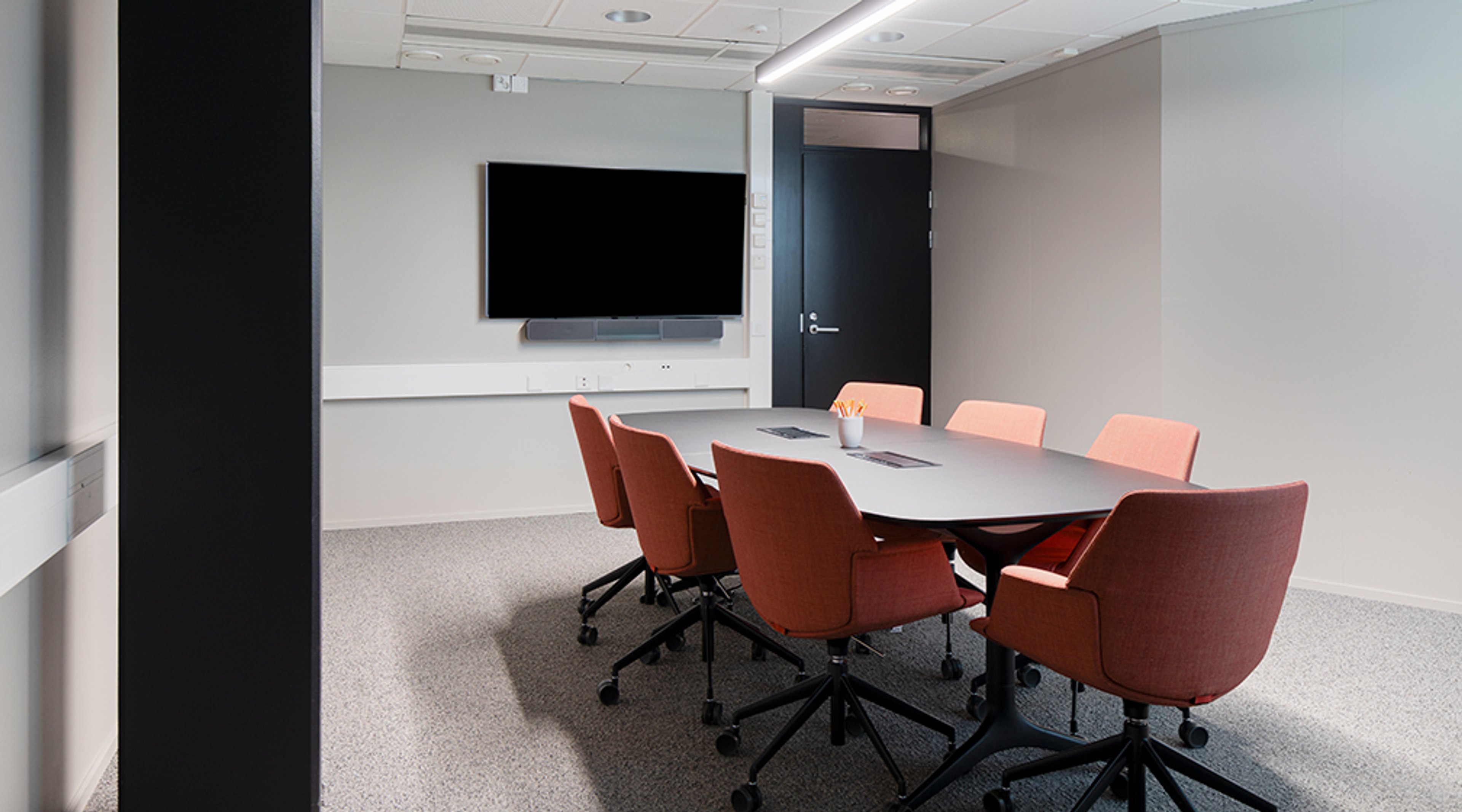 Moderni neuvotteluhuone, jossa soikea pöytä, terrakotanväriset pyörillä liikkuvat tuolit ja seinällä kiinnitetty näyttö. Huoneessa on kiinteä lattiapinta ja leveä oviaukko.