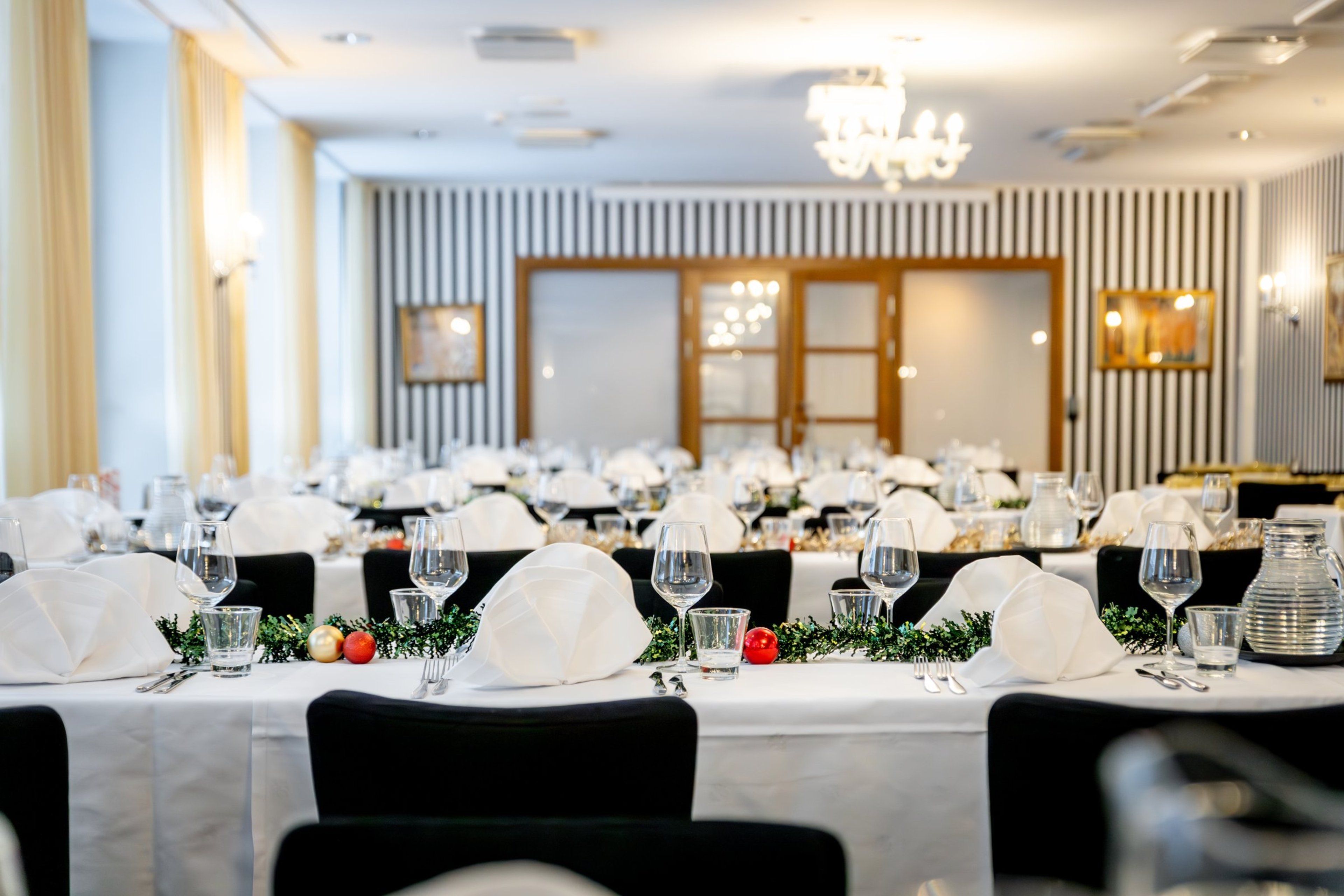 Juhlavasti katettu ruokasali valkoisilla pöytäliinoilla, mustilla tuoleilla ja joulukoristeilla pöydällä. Taustalla juhlasalin raidallinen seinä ja puiset ovet.