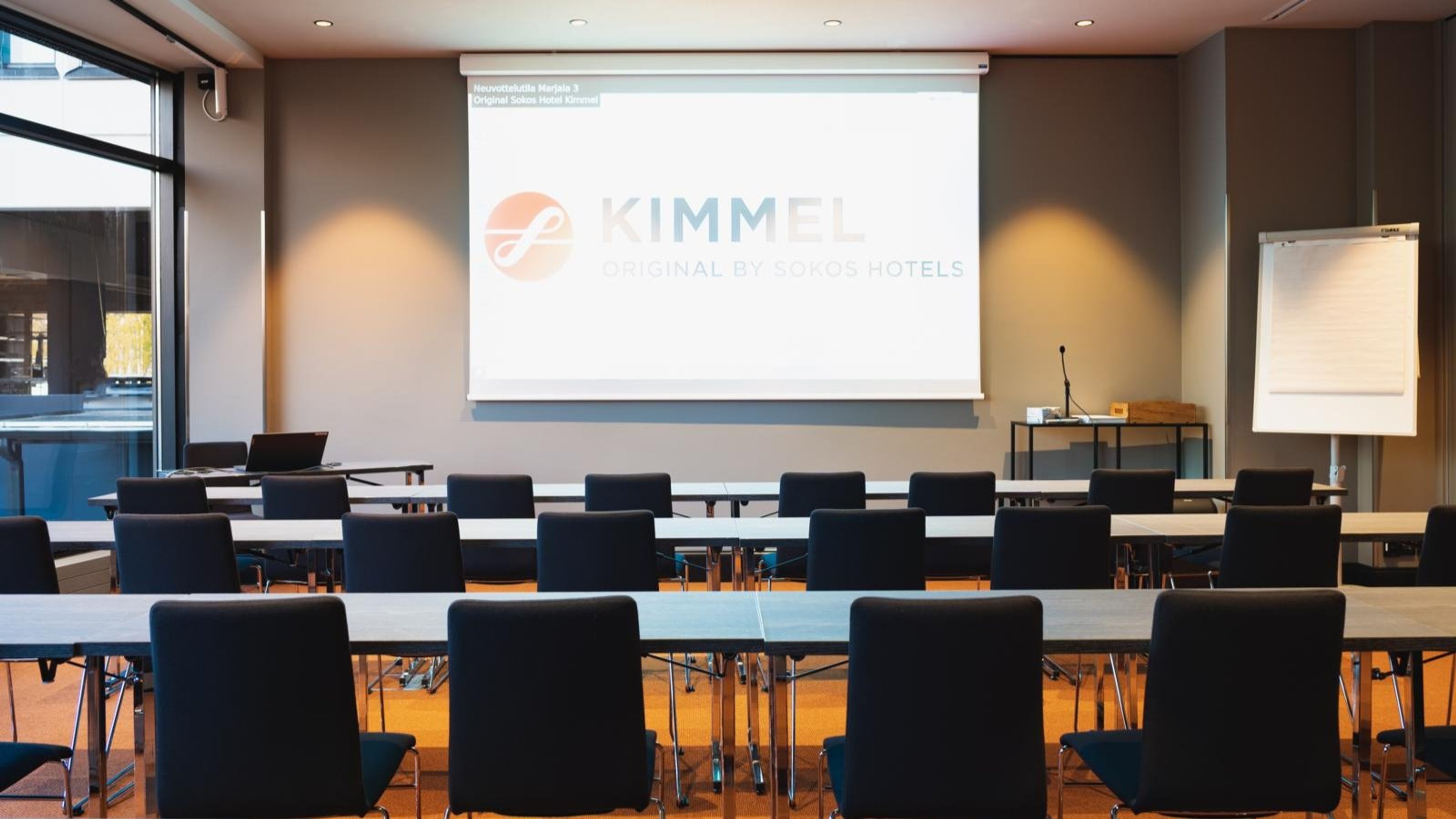 Kokoushuone pitkillä pöydillä, mustilla tuoleilla ja projektorikankaalla. Huoneessa suuret ikkunat ja tasainen lattia.