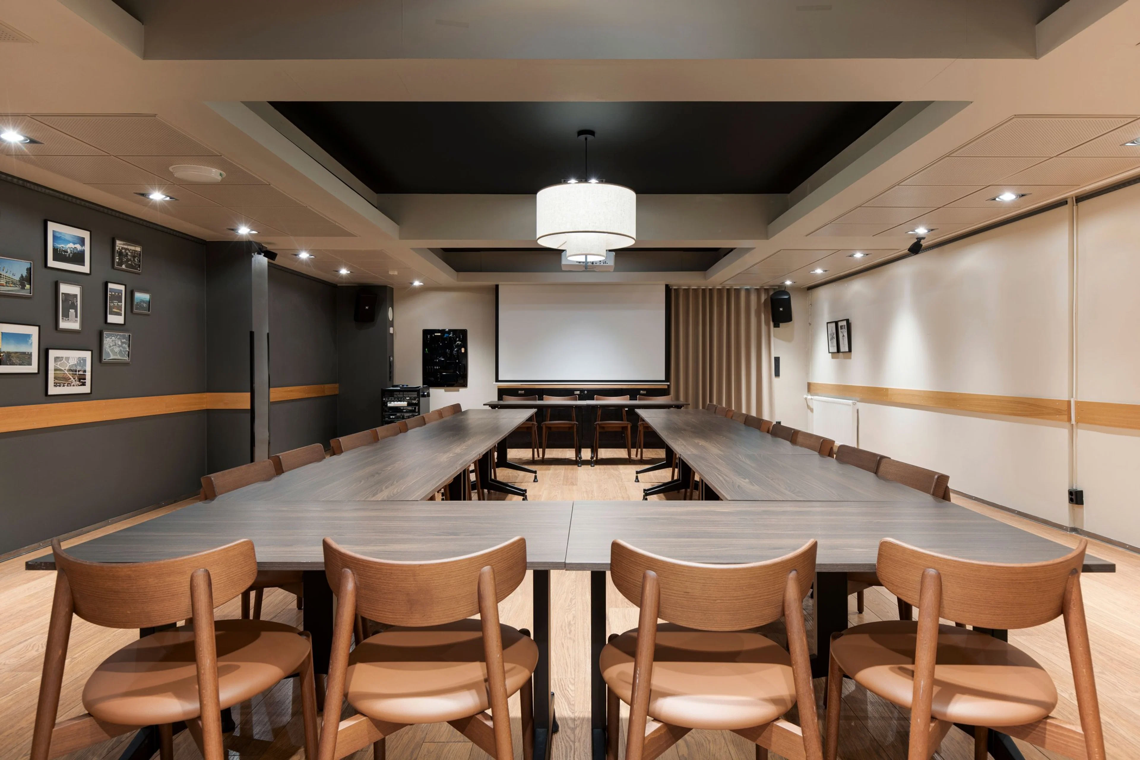 Moderni kokoushuone U-muotoisella pöytäjärjestelyllä, puiset tuolit, valkokangas edessä ja puulattia. Tumma ja vaalea seinäväritys.