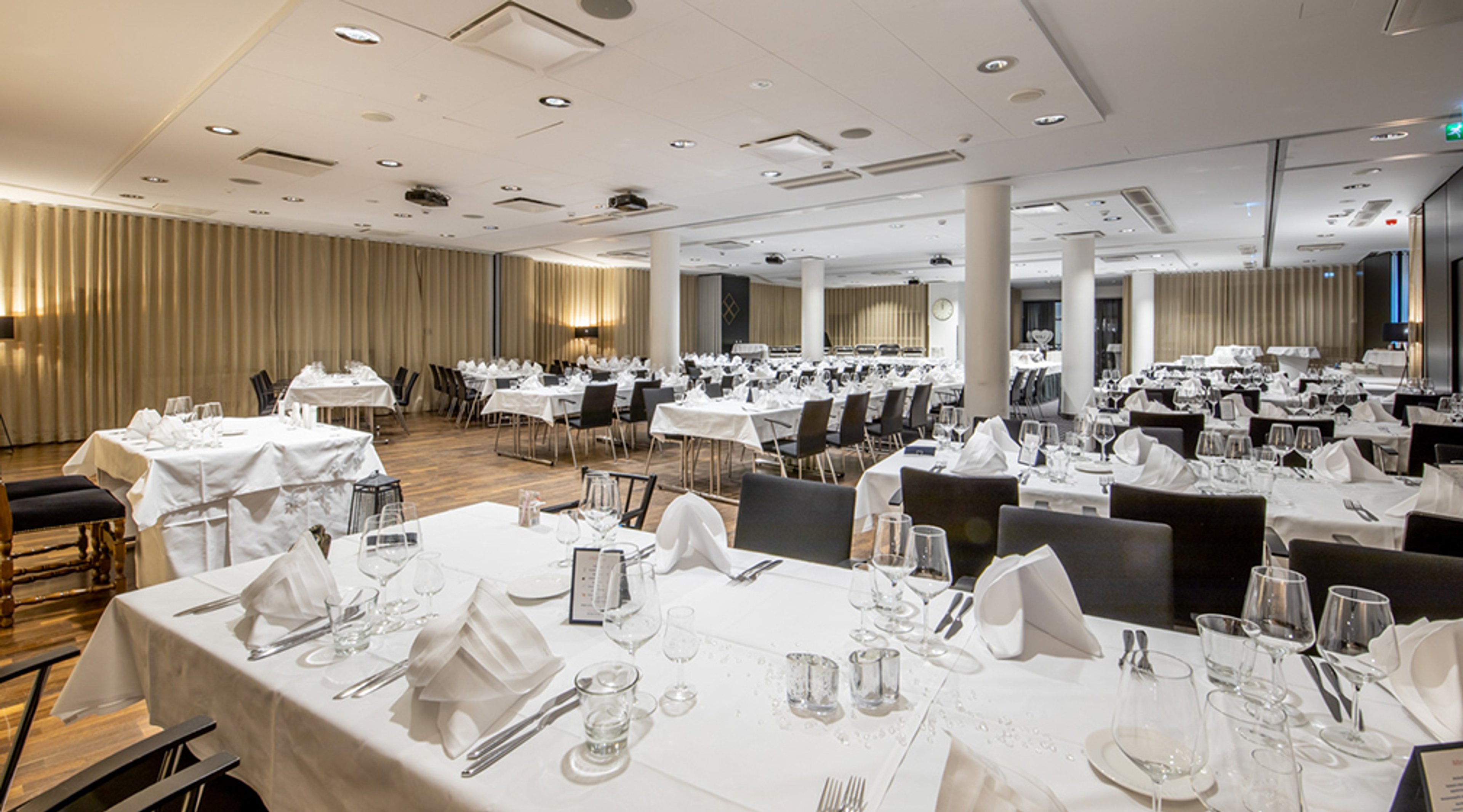 Avara juhlatila, jossa useita pöytiä valkoisten liinojen ja kattausten kera. Puulattia ja betonipylväät modernissa sisustuksessa.