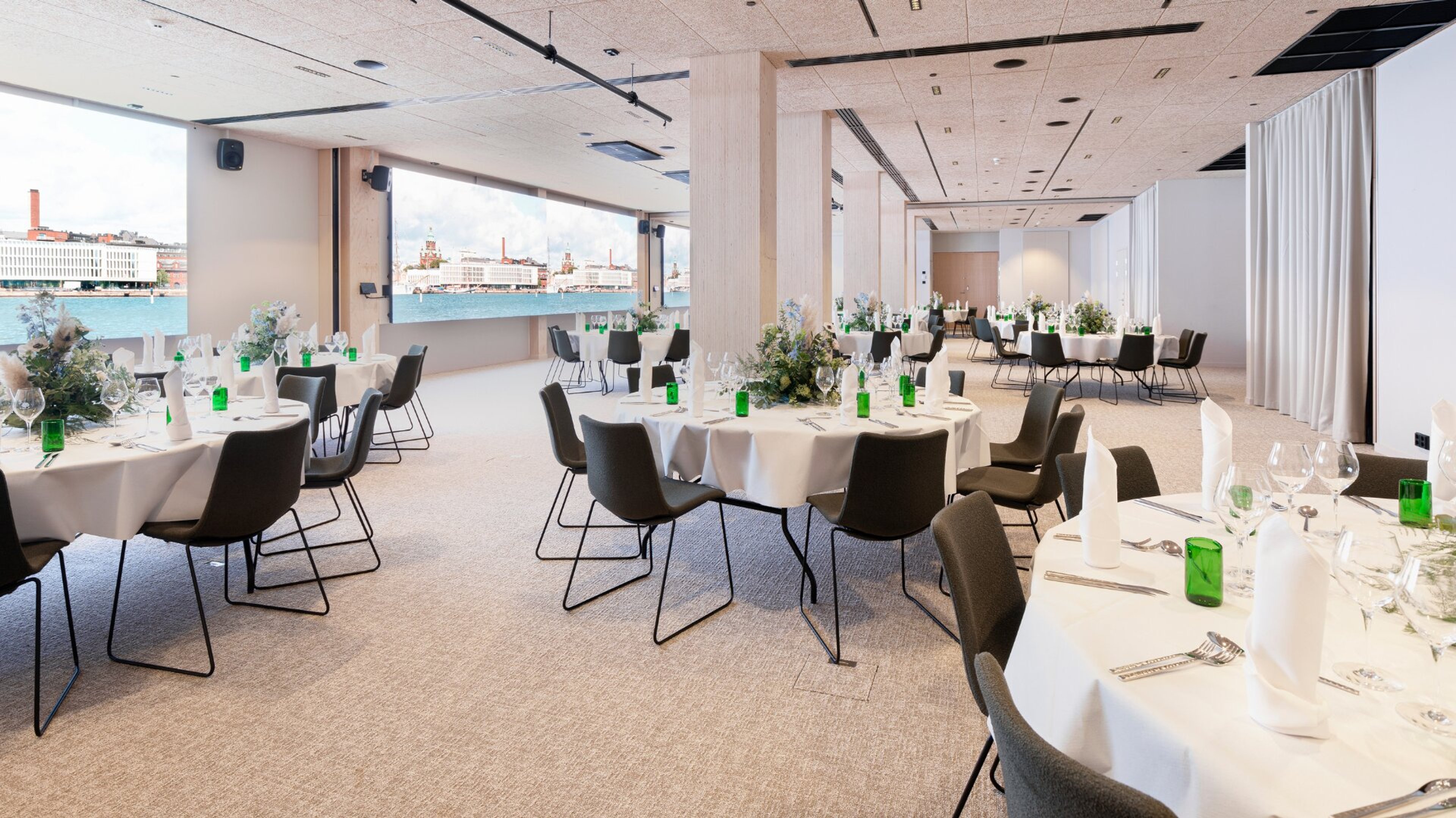 Modern festlokal med runda bord. Stort fönster med utsikt över stadens hamn. Matta på golvet och tillgängligt utrymme mellan borden.