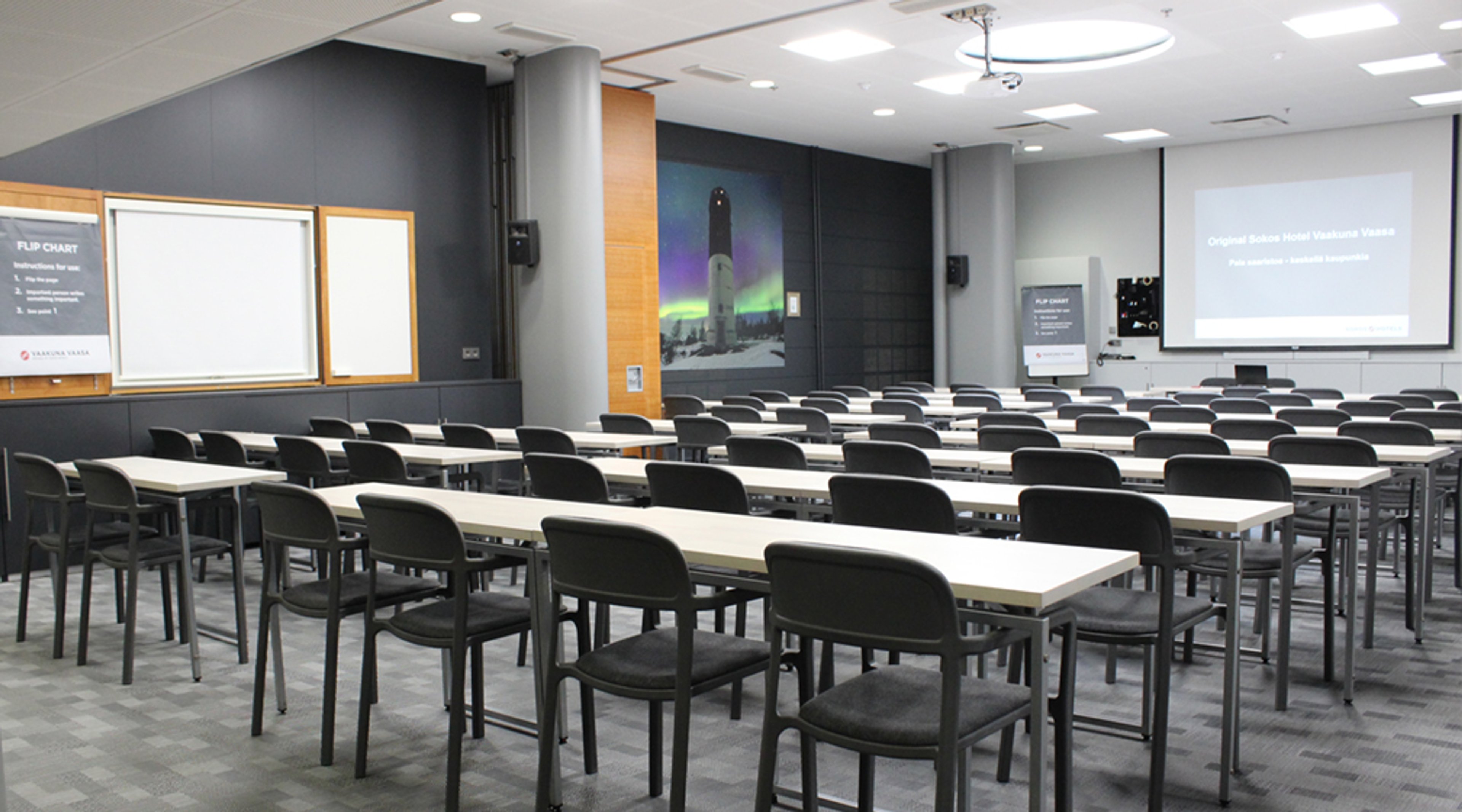 Luokkahuone pitkillä pöydillä ja tuoleilla, edessä valkokankaat ja näyttö. Harmaa-siniset seinät, joista yhdessä pohjoisen revontulikuva.