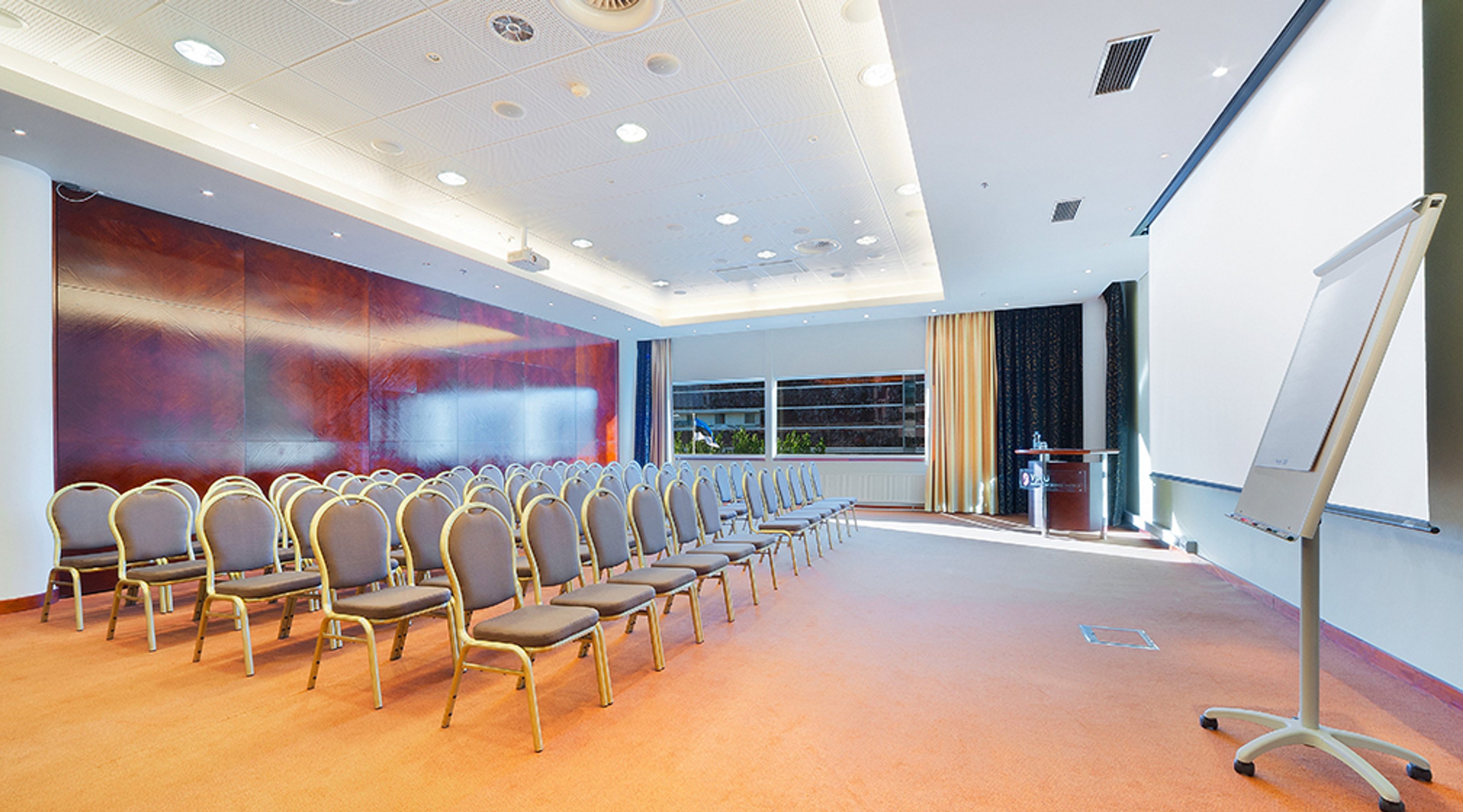 Moderni kokoushuone punaisella seinällä, riveittäin tuoleja, tasainen lattia ja valkotaulu esityksiä varten.