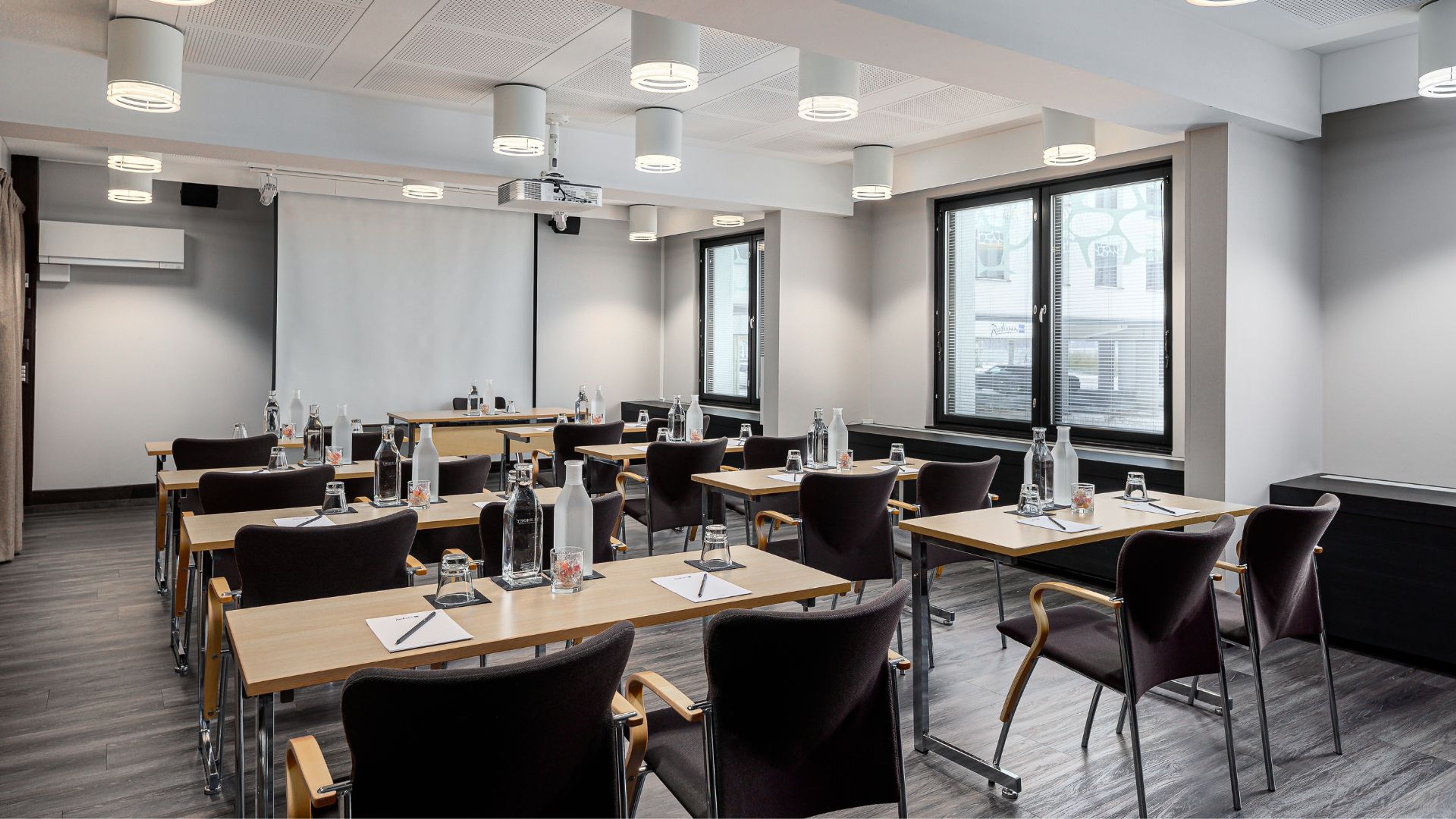 Moderni kokoustila puisilla pöydillä ja mustilla tuoleilla. Tilassa on tasainen lattia, iso valkokangas ja hyvä valaistus katossa.