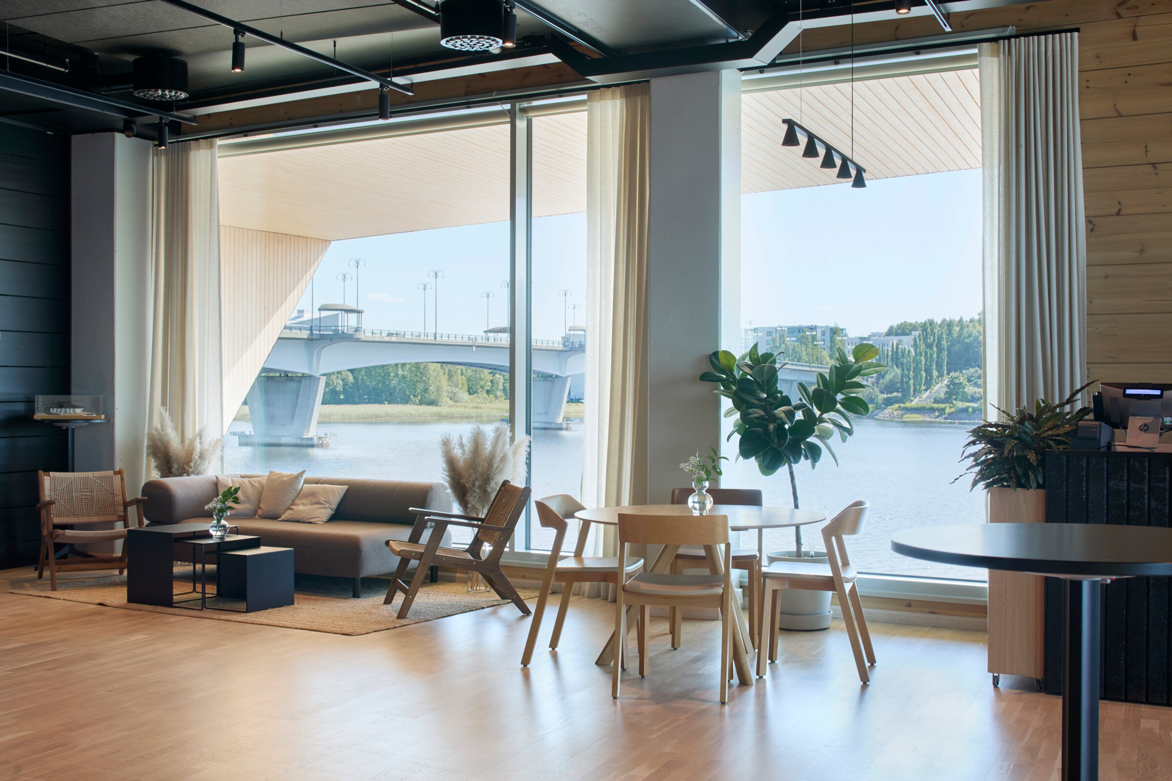 Moderni oleskelutila puulattialla, jossa on sohva, tuoleja ja ruokapöytä. Suurista ikkunoista avautuu näkymä joelle ja sillalle.